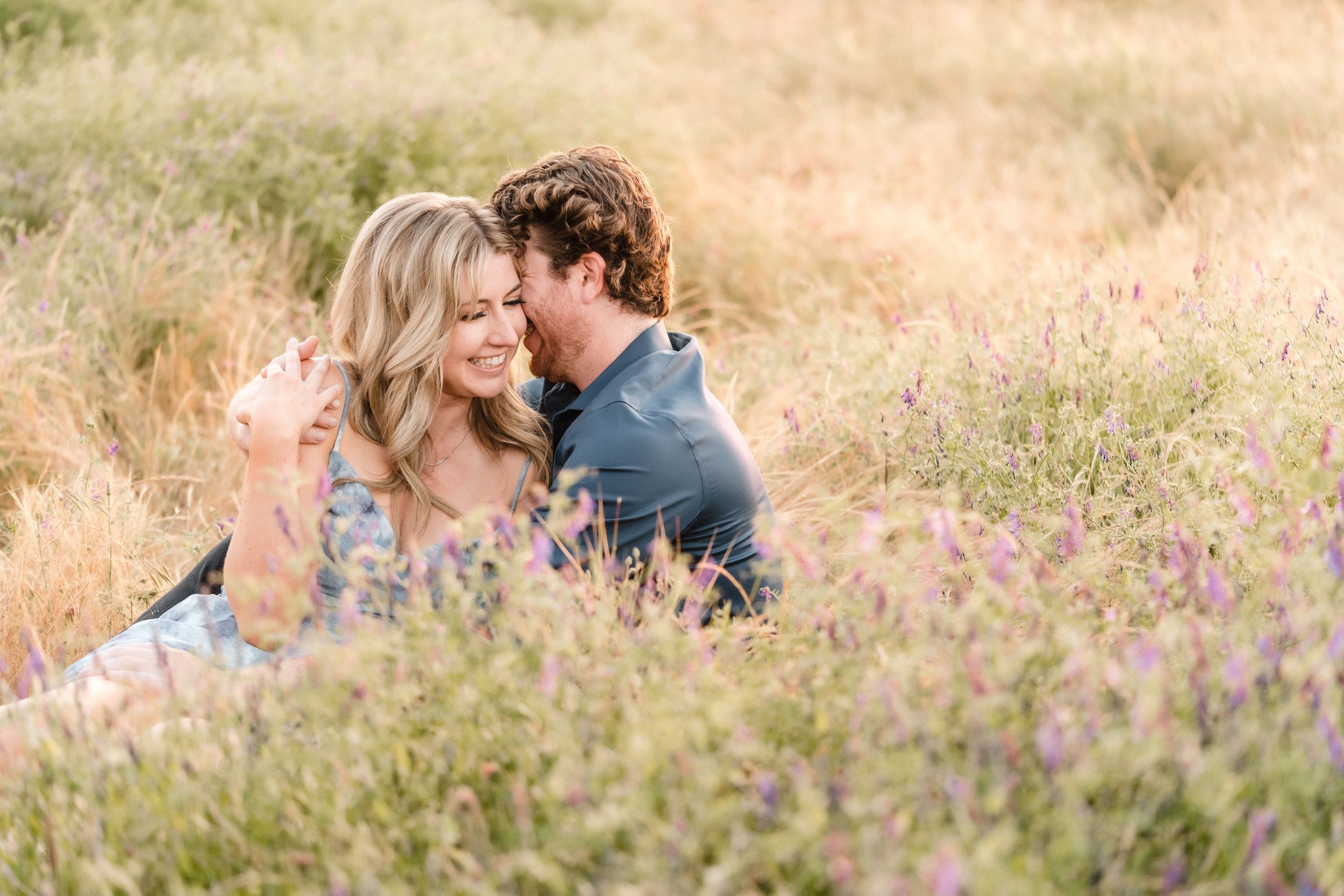 A happy couple with fair skin embracing in a field of wildflowers during sunset, smiling with their foreheads touching during their engagement session. Photography by Katherine Nicole Photography.