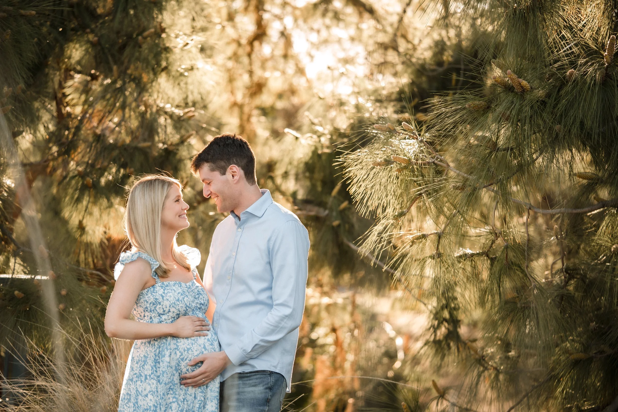A couple stands closely together in a forested area during sunset, smiling and looking into each other's eyes with trees and sunlight in the background, during a maternity session with Sacramento family photographer Katherine Nicole Photography. 