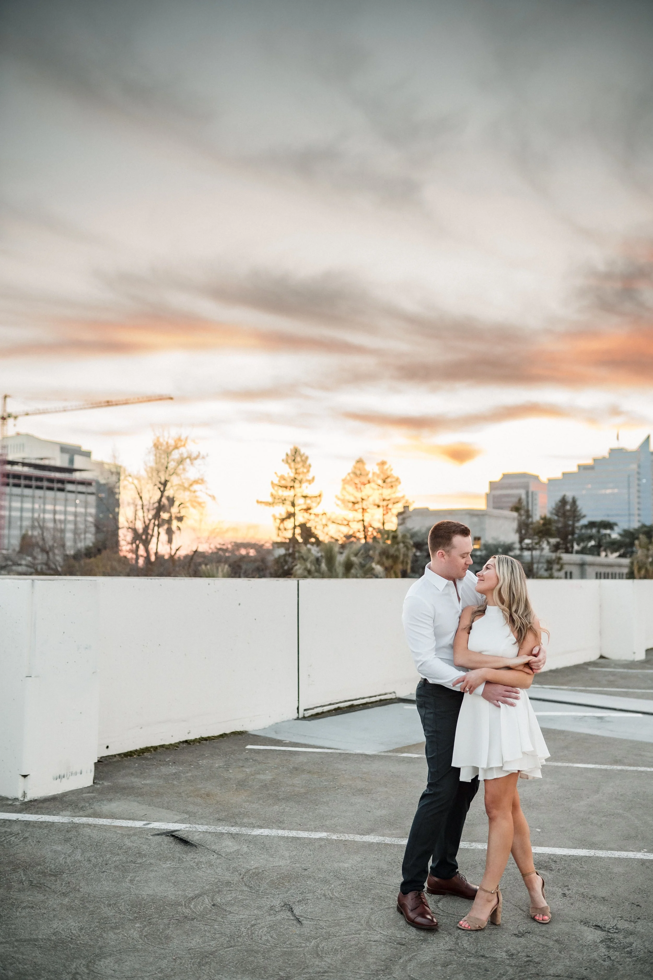 A couple gazes lovingly at each other on a Sacramento rooftop parking lot at sunset during their engagement session, with a city skyline and cloudy sky in the background. Photography by Katherine Nicole Photography.