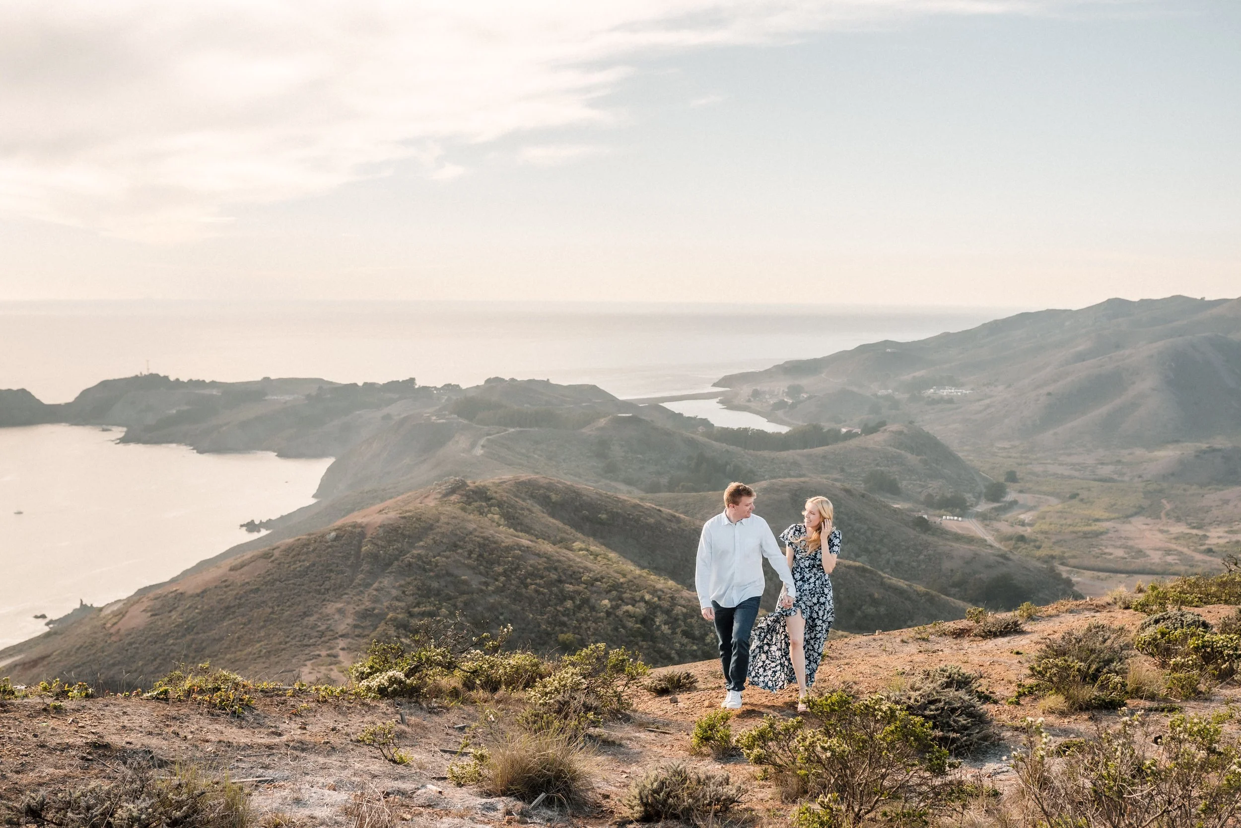A couple walking together on a hilltop overlooking a scenic landscape of hills, water, and distant coastline on a partly cloudy day during their Bay Area engagement session. Photography by Katherine Nicole Photography.