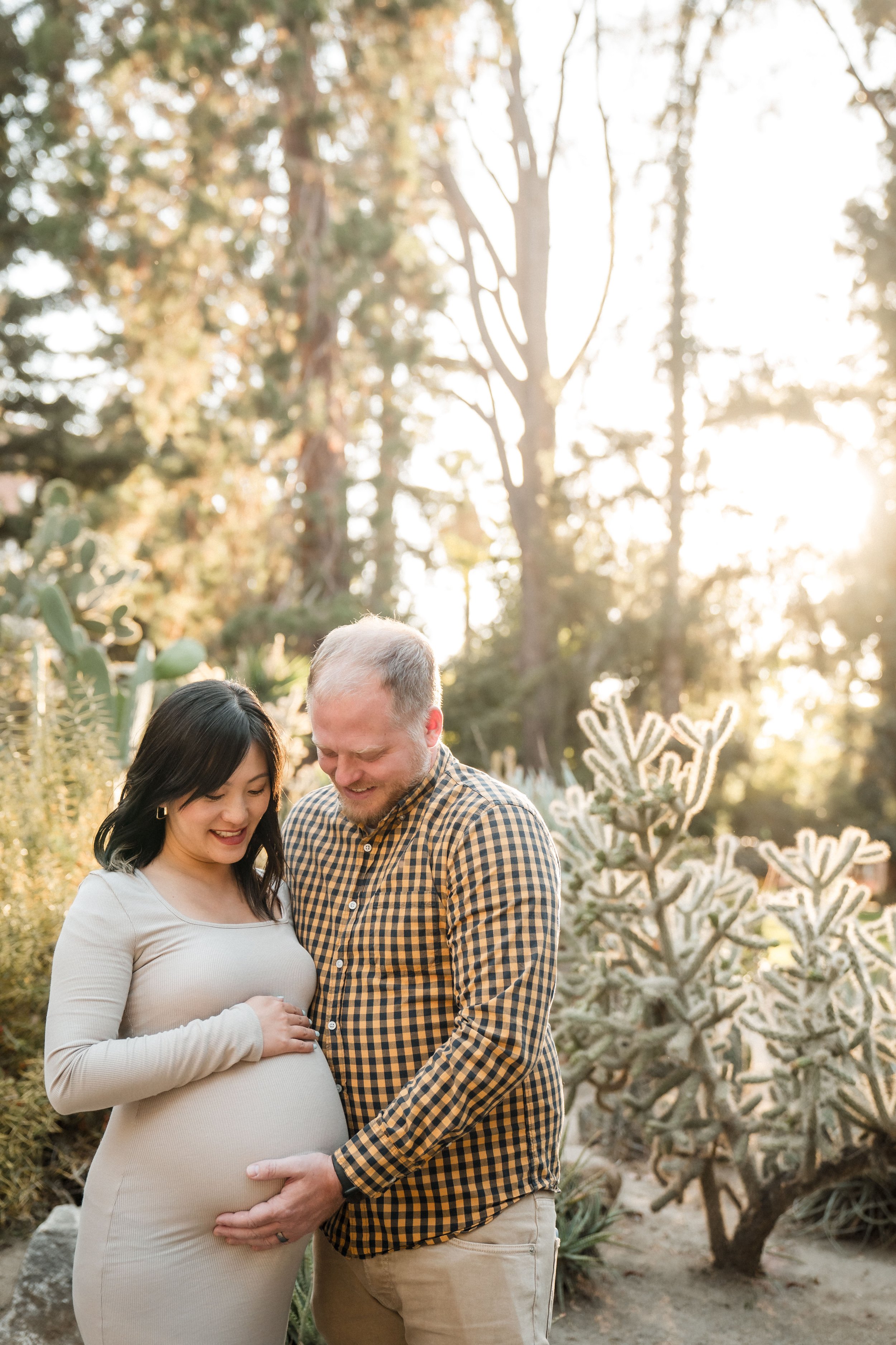 A pregnant woman with her husband, standing outdoors among trees and succulents, smiling and looking at her belly as she holds her stomach, during their maternity session with Sacramento family photographer Katherine Nicole Photography. 