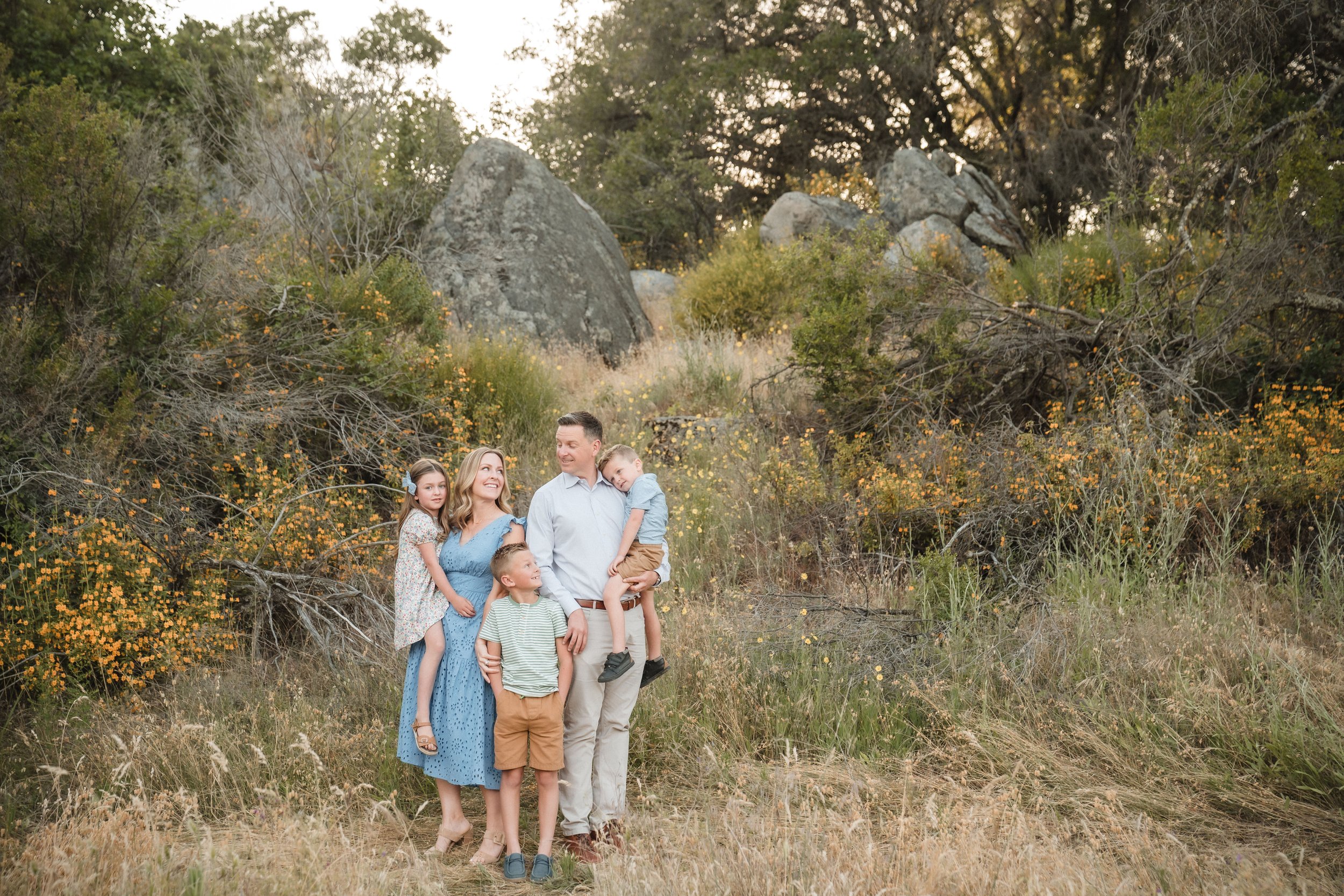 A family standing outdoors in a grassy area with trees and large rocks in the background, looking at each other, during their family photos at Folsom Lake with Sacramento family photographer, Katherine Nicole Photography.