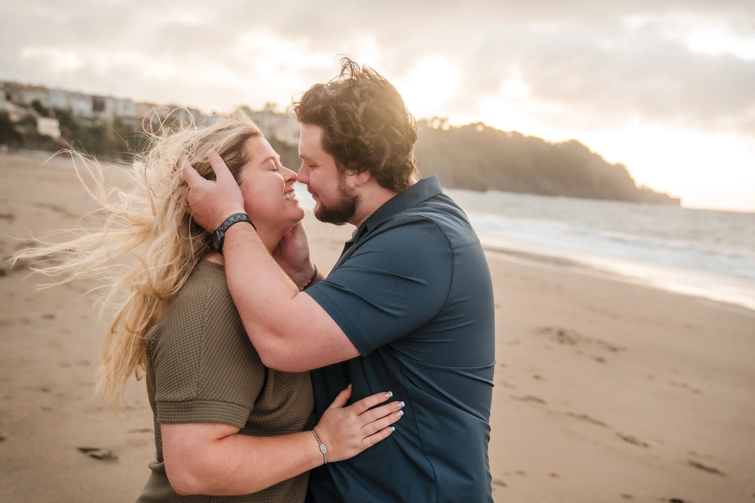 A man and woman on a beach, close to each other, smiling and touching foreheads during their Bay Area engagement session, with beach houses and cloudy sky in the background. Photography by Katherine Nicole Photography.