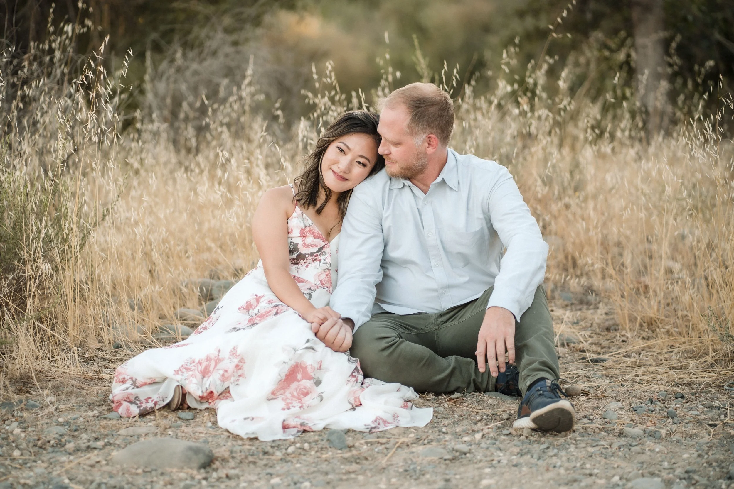 A couple sitting closely together outdoors in a field of dry grass, with the woman leaning her head on the man's shoulder during their engagement photos in Sacramento, CA. Photography by Katherine Nicole Photography.
