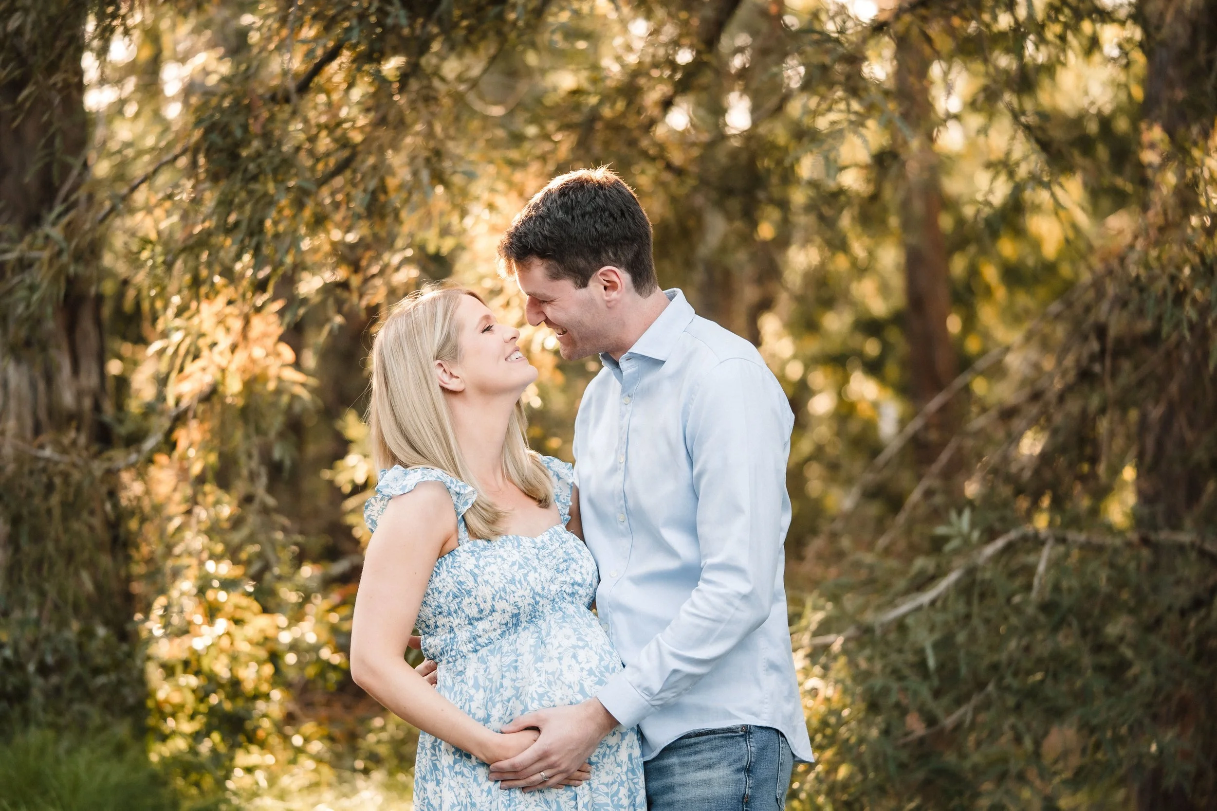 A couple, taking maternity photos with Katherine Nicole Photography at the UC Davis arboretum,  standing close to each other outdoors in a forested area with sunlight filtering through trees, smiling and looking into each other's eyes.