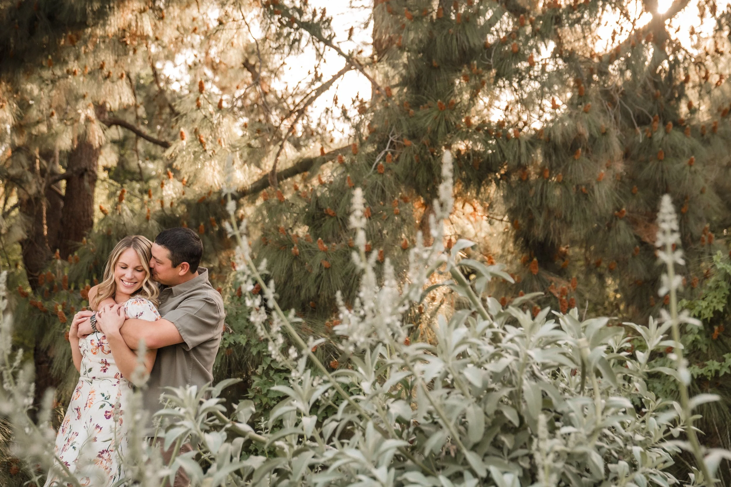 A smiling woman and man embrace in a lush garden with tall pine trees at their UC Davis Arboretum engagement session with Katherine Nicole Photography.