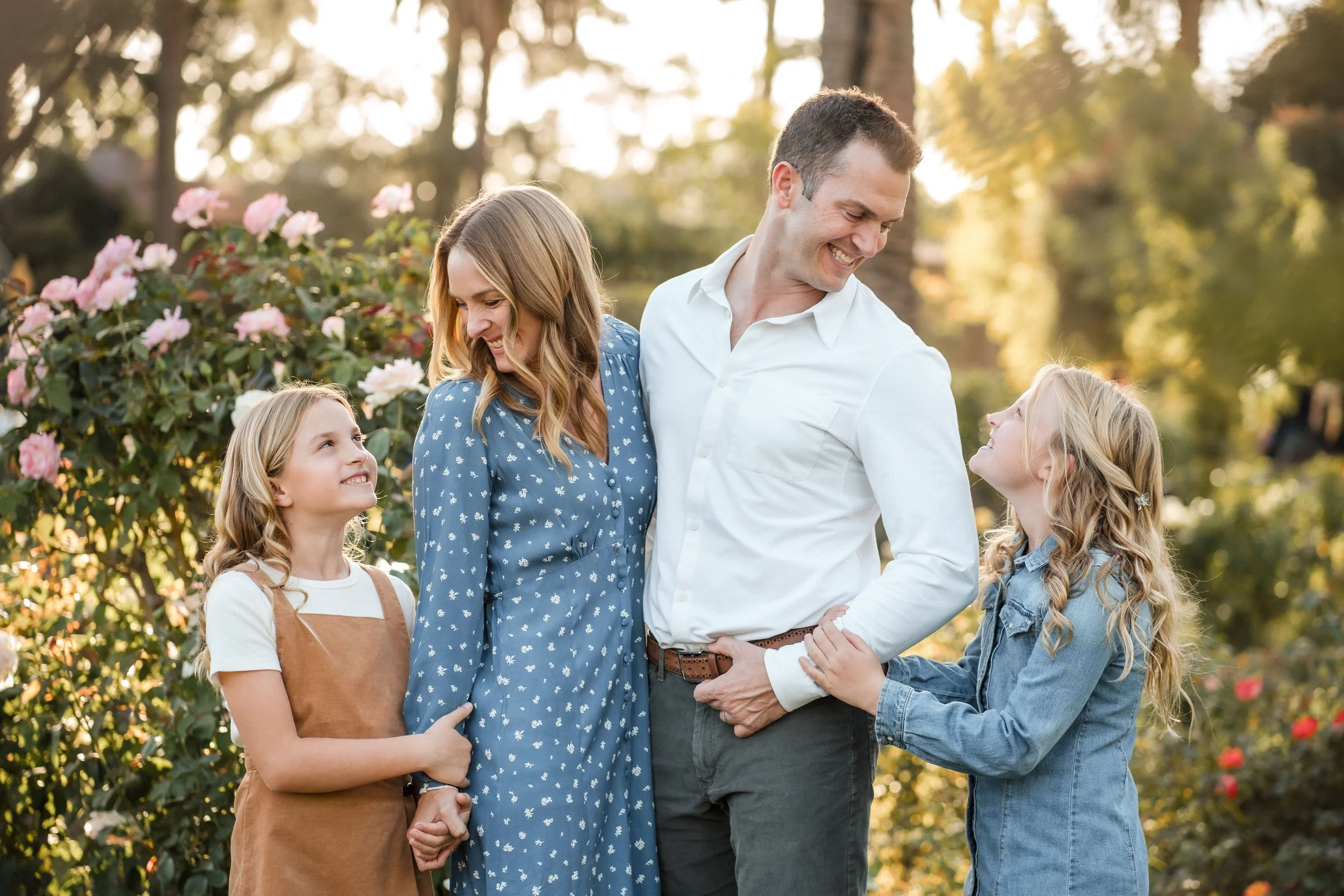 A smiling family of four, with two young girls, enjoying a walk outdoors in a park with blooming pink flowers and trees in the background during family photos with Sacramento family photographer, Katherine Nicole Photography.