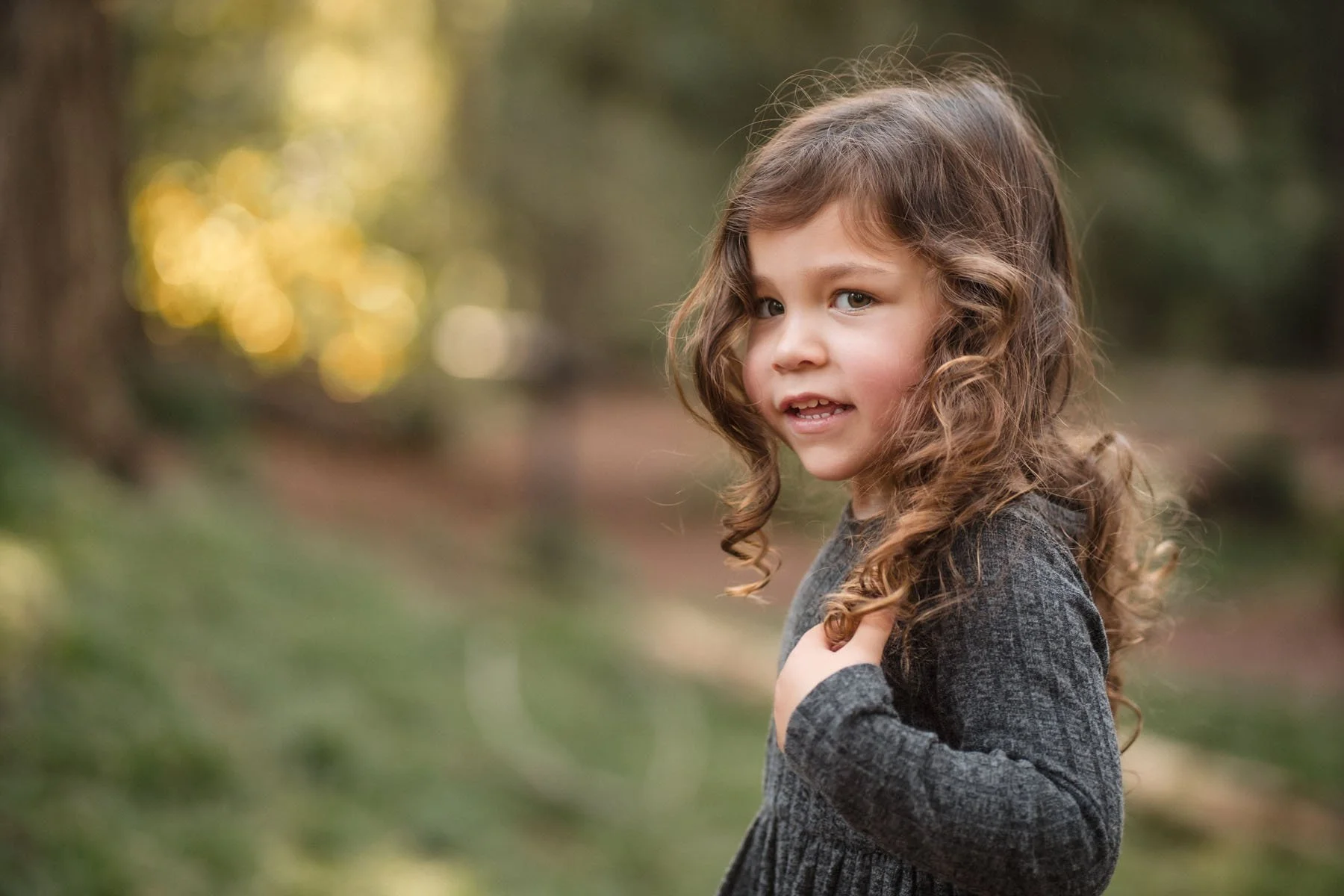 A young girl with long, curly brown hair and light skin standing outdoors in a park during autumn, wearing a gray sweater, with a blurred background of trees and foliage.