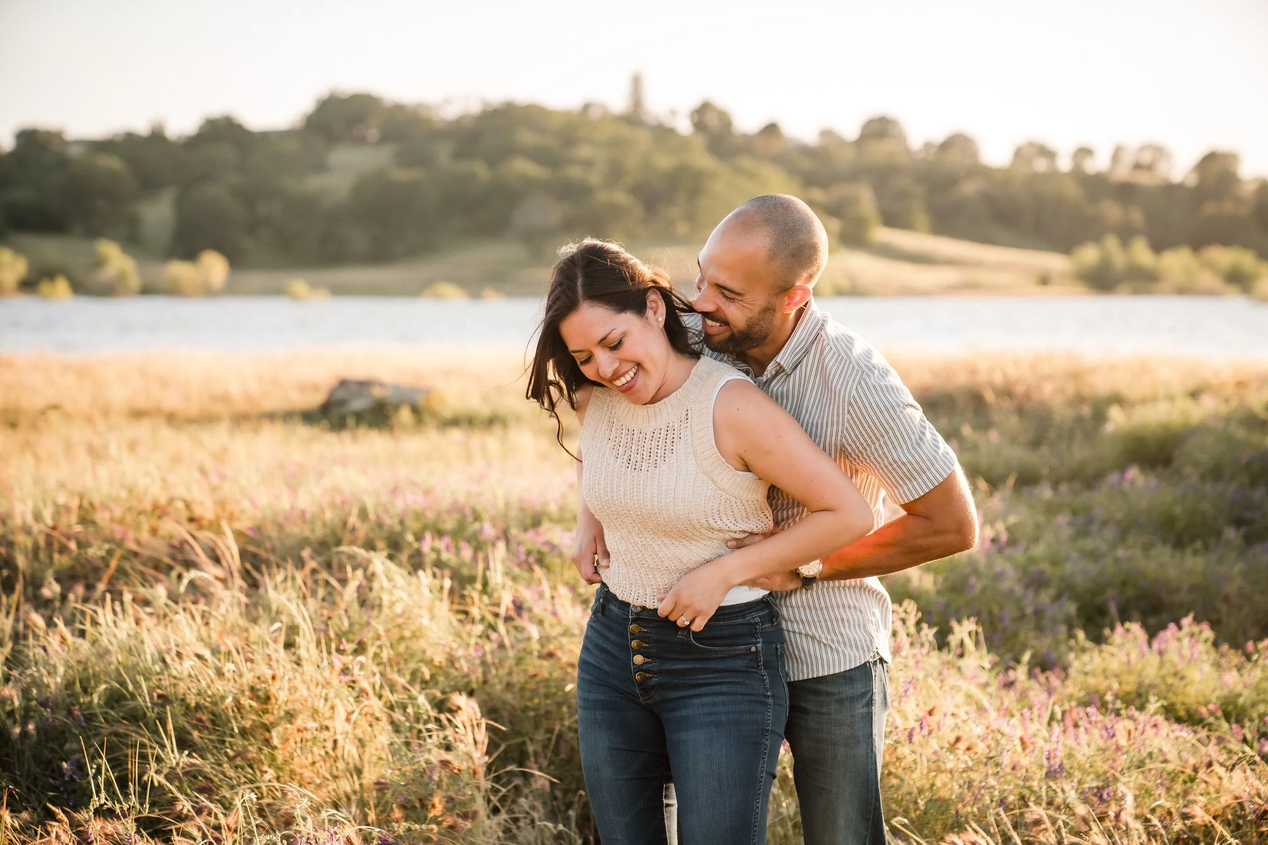 A smiling couple embracing in a field of wildflowers during sunset with Folsom Lake and rolling hills in the background, during their engagement session with Katherine Nicole Photography.