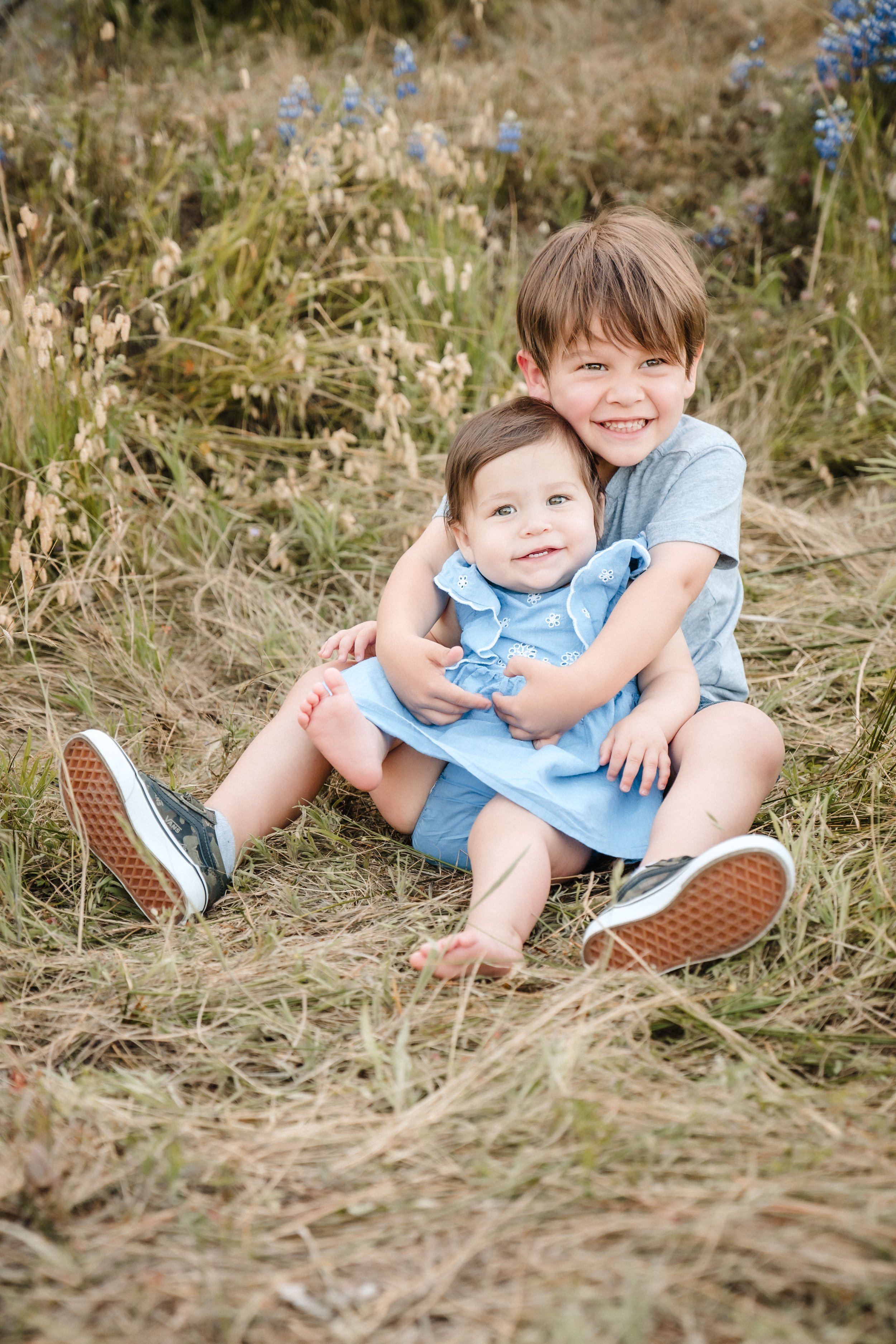 Two young children, a boy and a girl, sitting on the grass outdoors among dry grass and wildflowers, smiling and hugging each other during family photos with Sacramento family photographer Katherine Nicole Photography. 