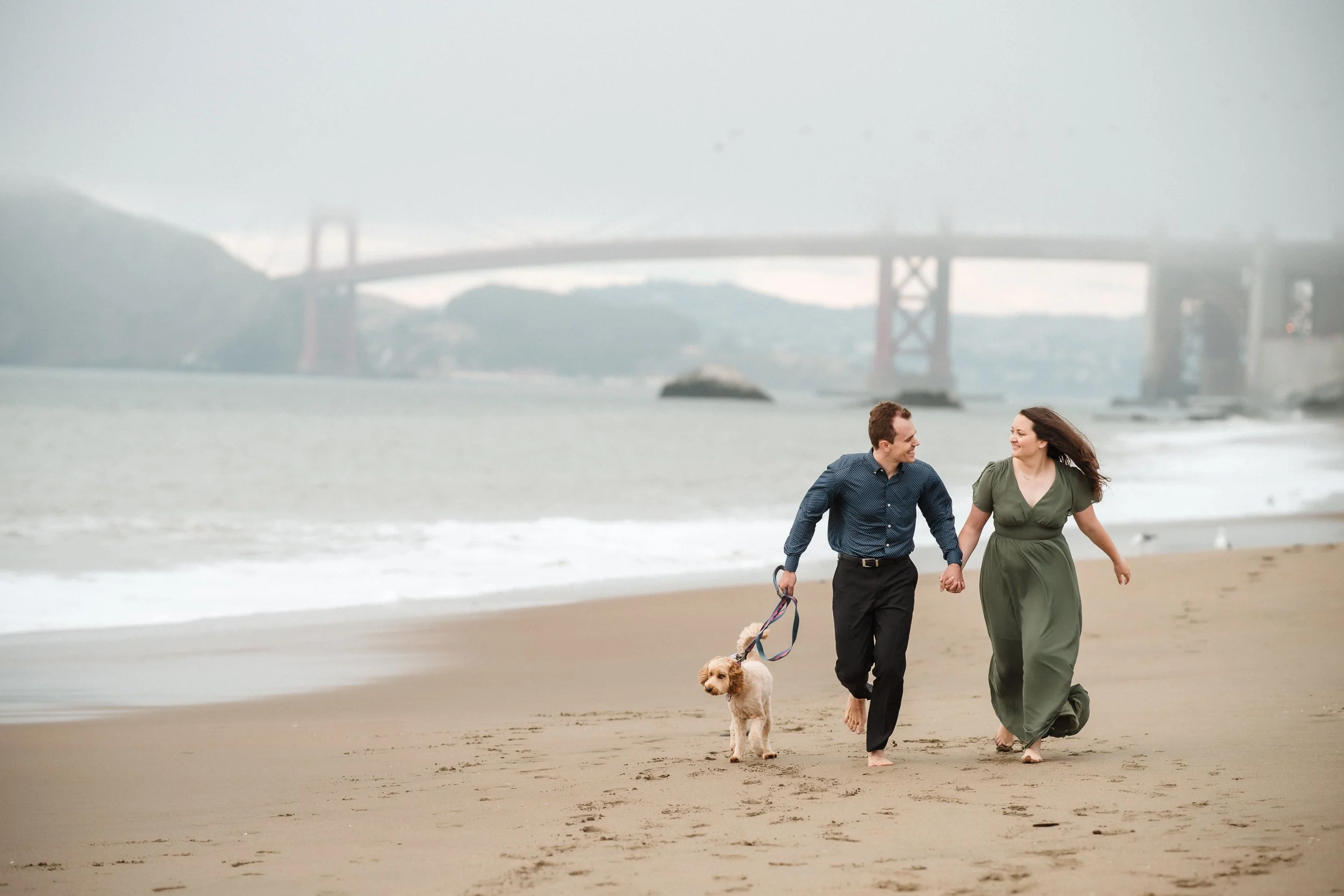 A couple walking and holding hands on a beach with their dog during their Bay Area engagement session. The background shows the Golden Gate Bridge and a cloudy sky. Photography by Katherine Nicole Photography.