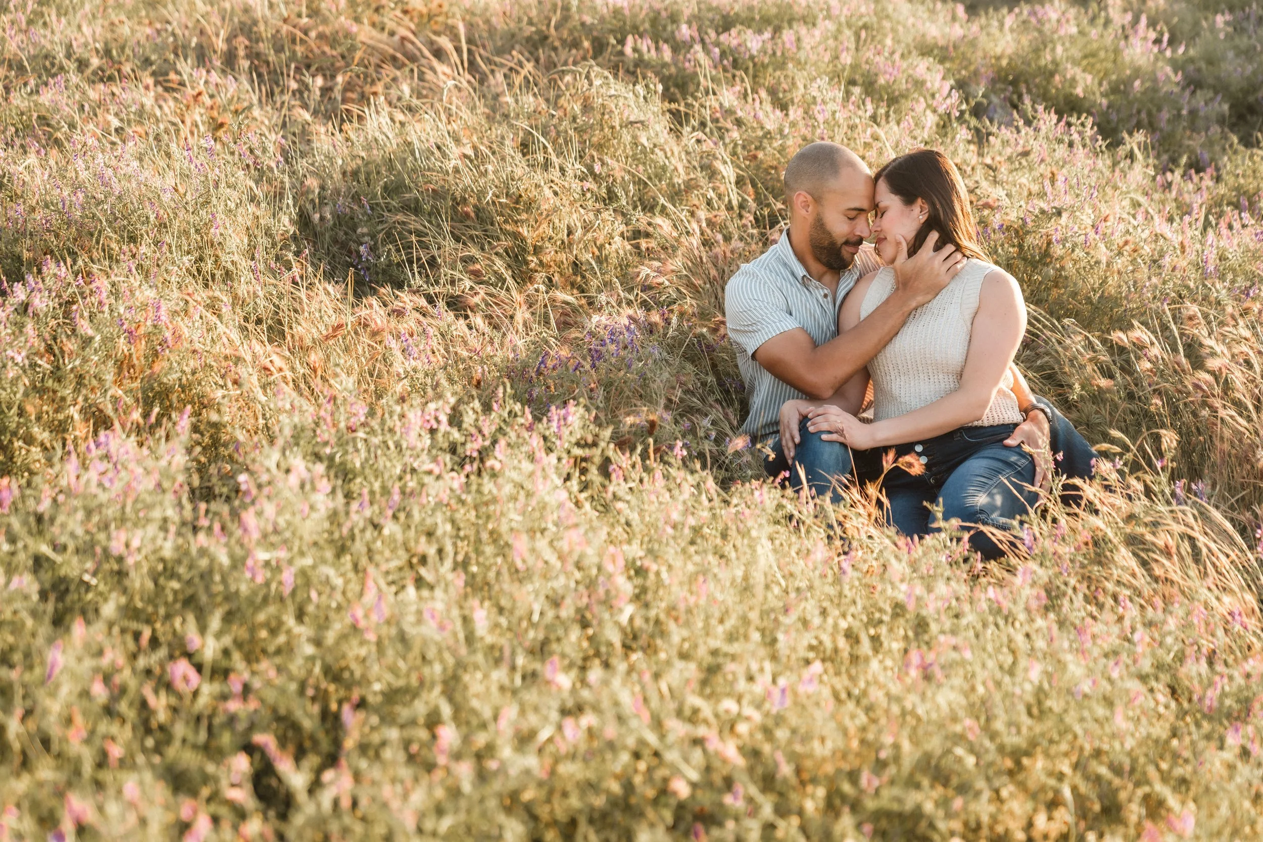 A couple sitting in a field of mixed wildflowers, touching foreheads and embracing in the sunlight during their Folsom Lake engagement session. Photography by Katherine Nicole Photography.