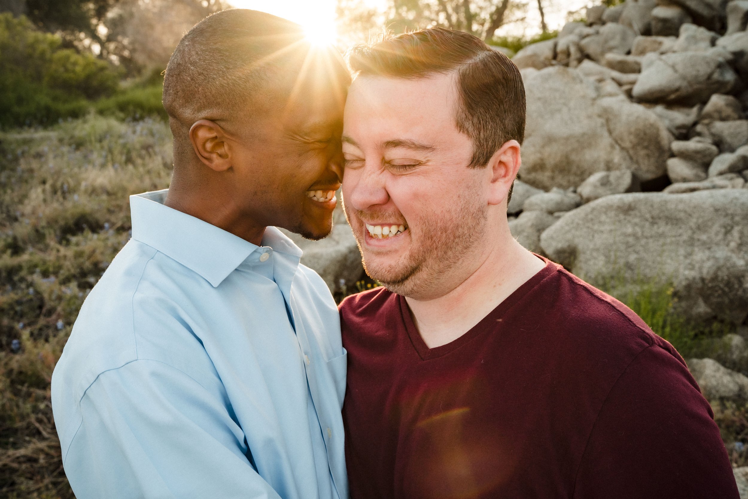 Two men smiling and pressing their foreheads together at their engagement session during sunset, with rocks and trees in the background. Photography by Katherine Nicole Photography.