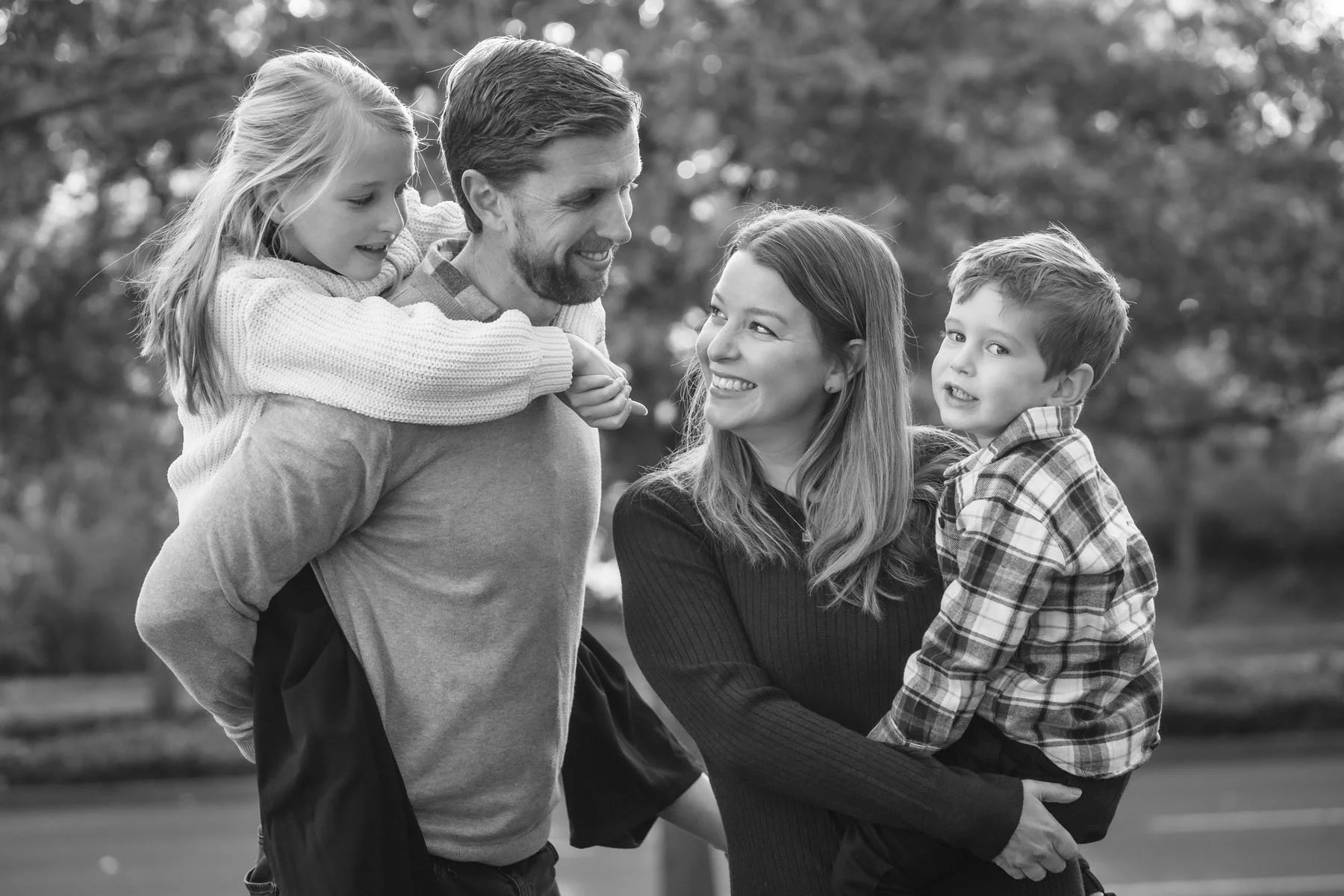 A black-and-white photo of a family outdoors, with a man carrying a young girl on his back, a woman holding a young boy, all smiling and looking at each other.