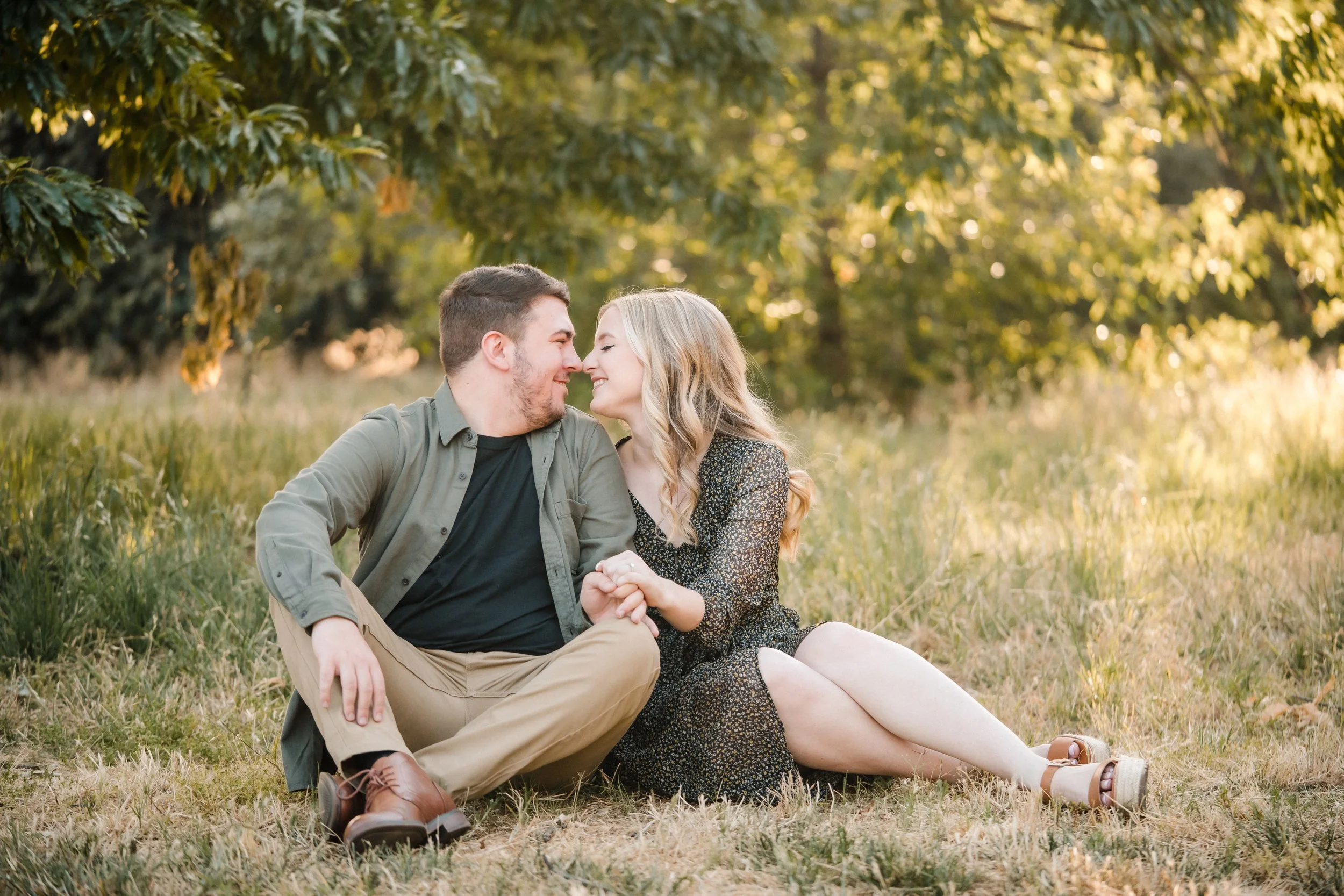 A young couple sitting on the grass during their UC Davis Arboretum engagement session, smiling and touching foreheads, with trees in the background. Photography by Katherine Nicole Photography.