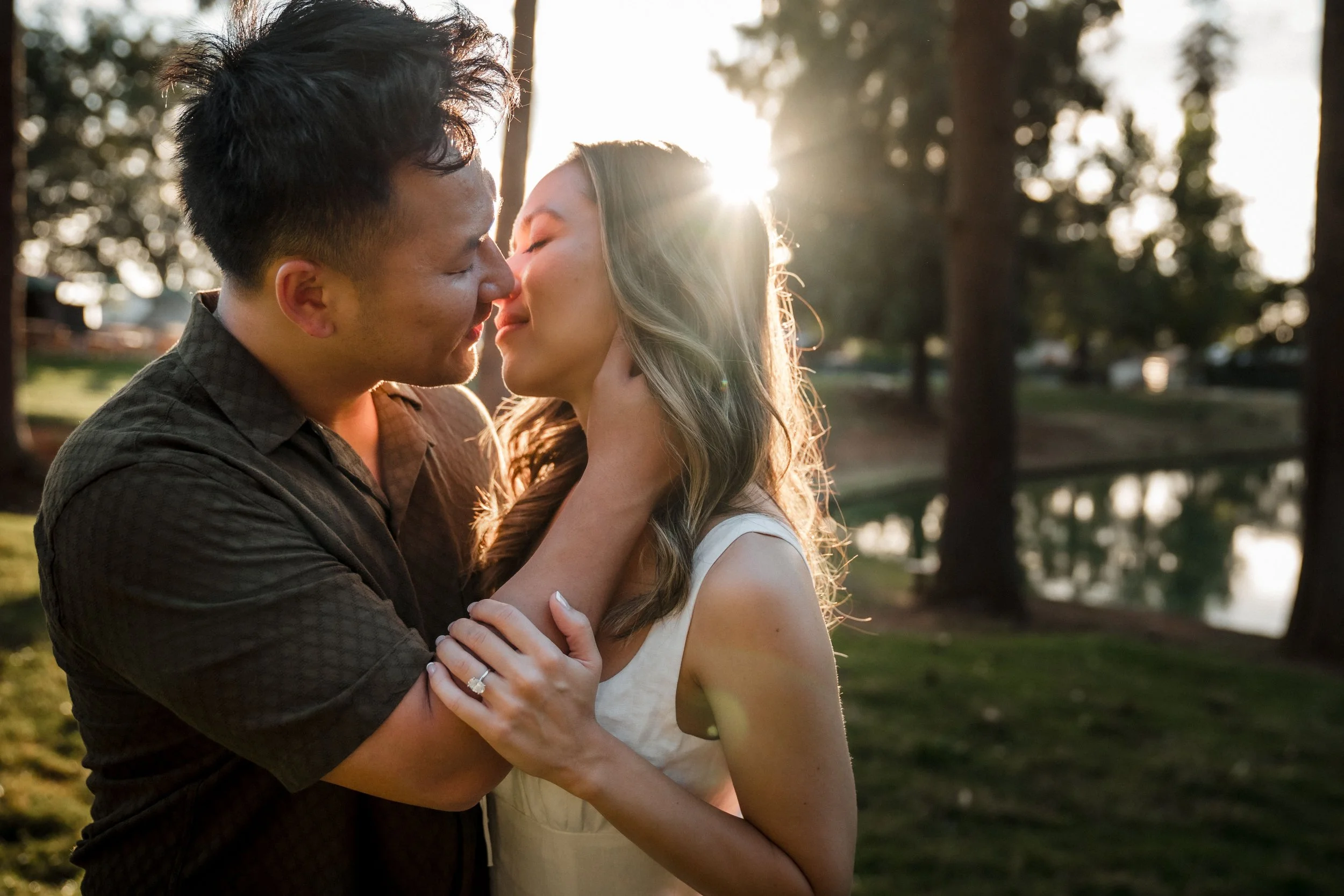 A couple kissing outdoors at sunset, embracing each other closely in front of tall trees with sunlight filtering through during their engagement session in Apple Hill with Katherine Nicole Photography.