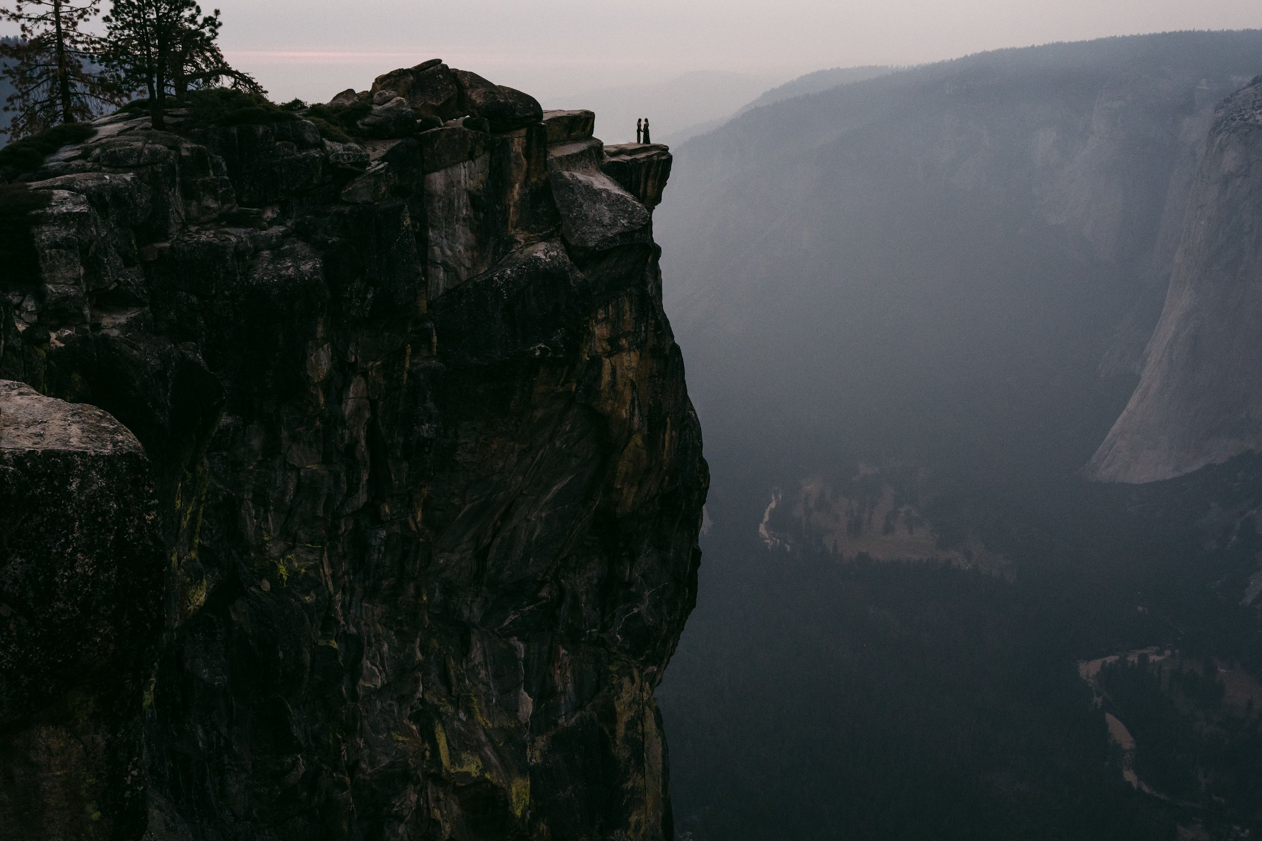 Two people standing on the edge of a rocky cliff at Taft Point in Yosemite with fog and mountain ranges in the distance at dusk. Photography by Katherine Nicole Photography.