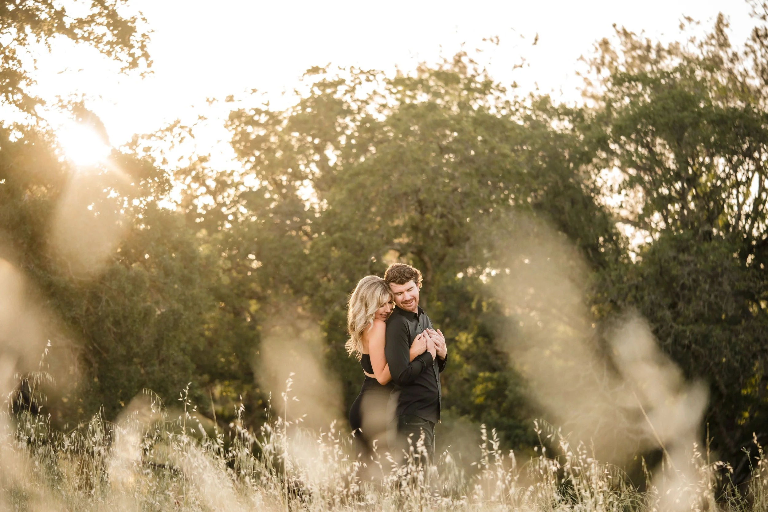 A couple embracing in a grassy field t sunset during their engagement session. Photography by Katherine Nicole Photography.