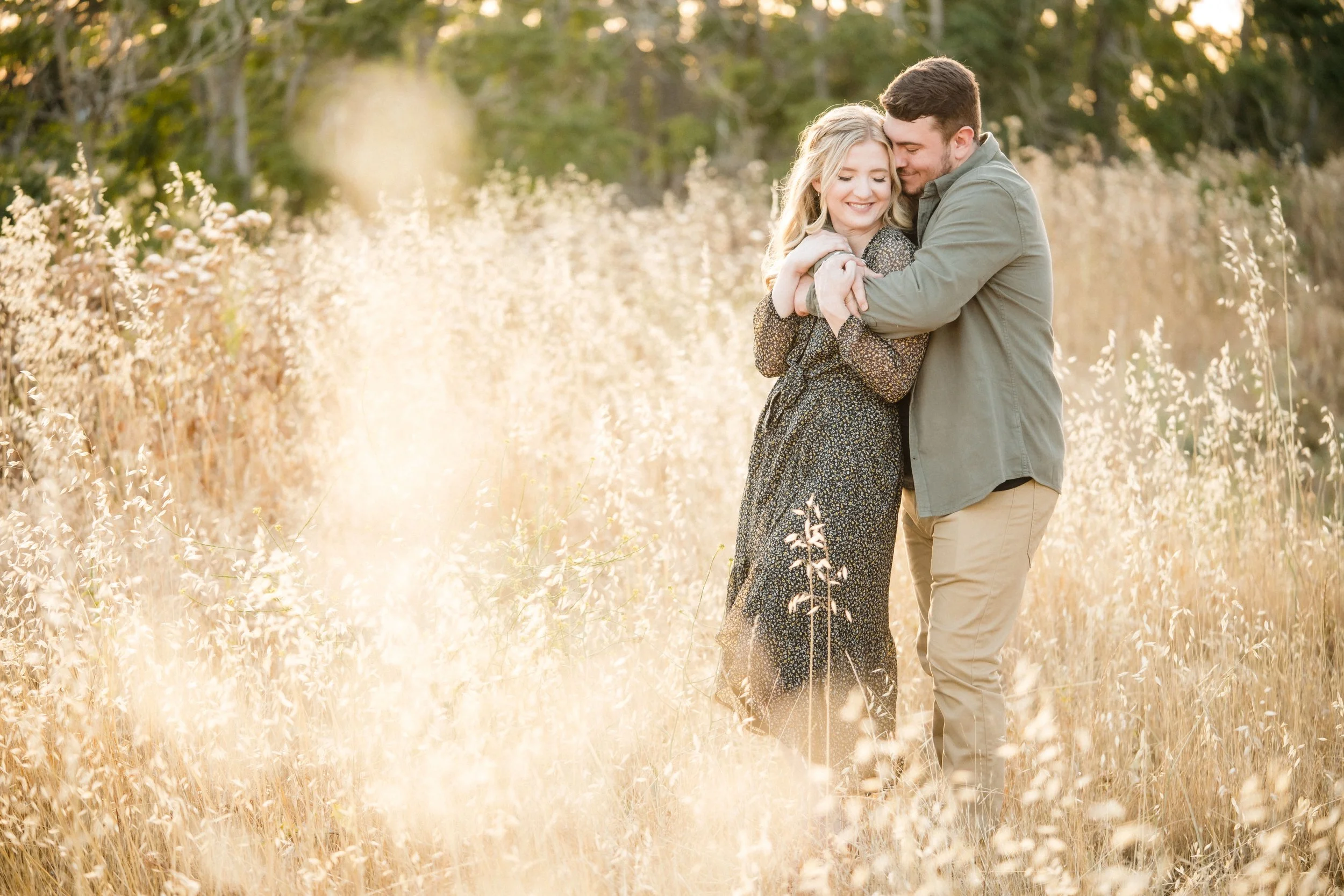 A young couple embracing in a field of tall, golden grass with trees in the background, during a warm sunset at their engagement session. Photography by Katherine Nicole Photography.