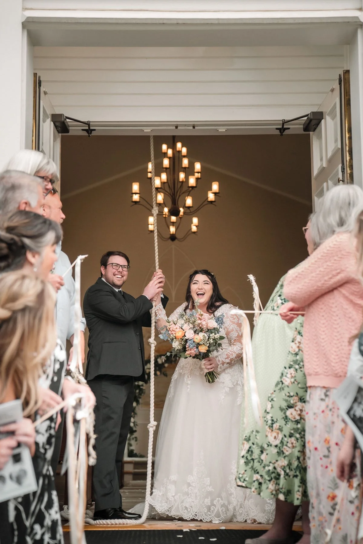 A wedding scene with a bride and groom holding a rope swing, surrounded by smiling guests, inside a church with a chandelier in the background, photographed by Sacramento wedding photographer Katherine Nicole Photography. 