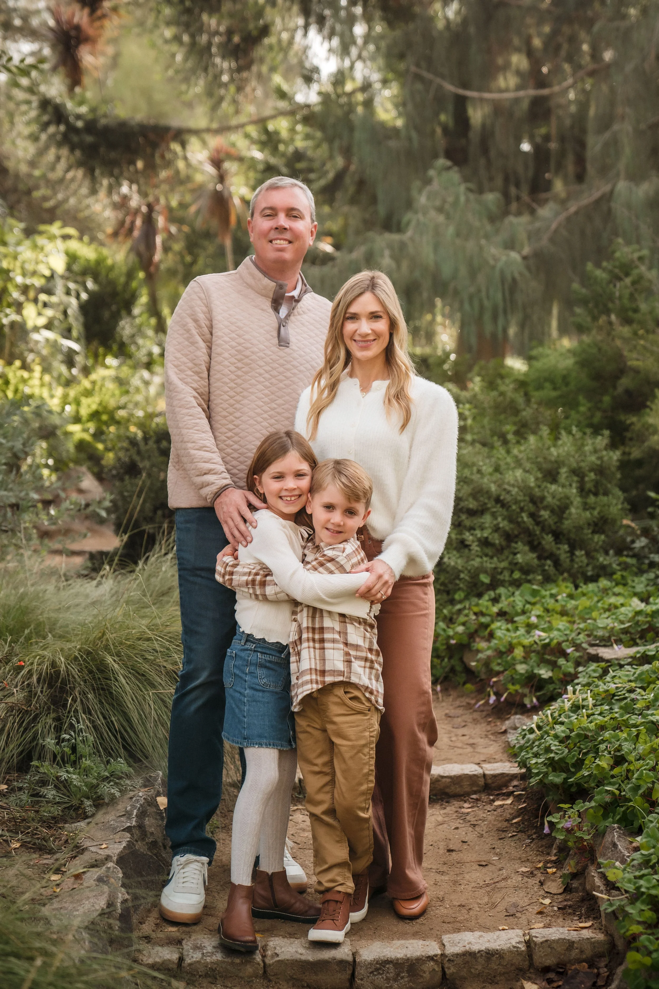 A family of four standing on a stone-paved path at the WPA Rock Garden, during their family photos with Katherine Nicole Photography, with trees and plants in the background, smiling at the camera.