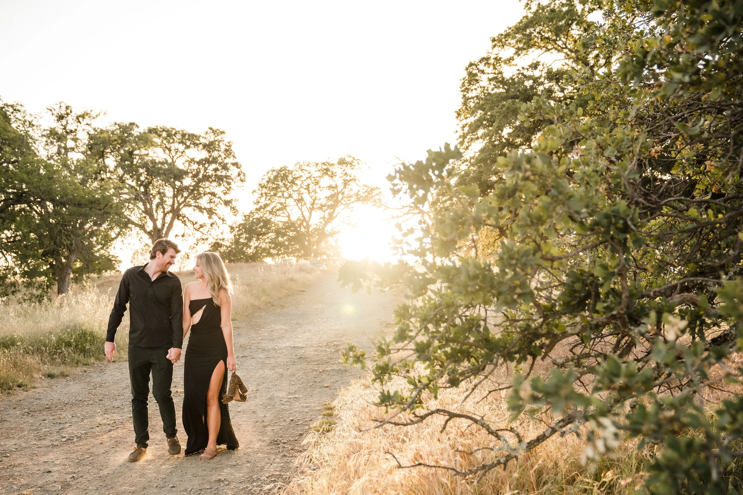 A young couple walking hand-in-hand on a dirt path during their engagement photos in a natural setting during sunset, with trees and foliage around them. Photography by Katherine Nicole Photography.