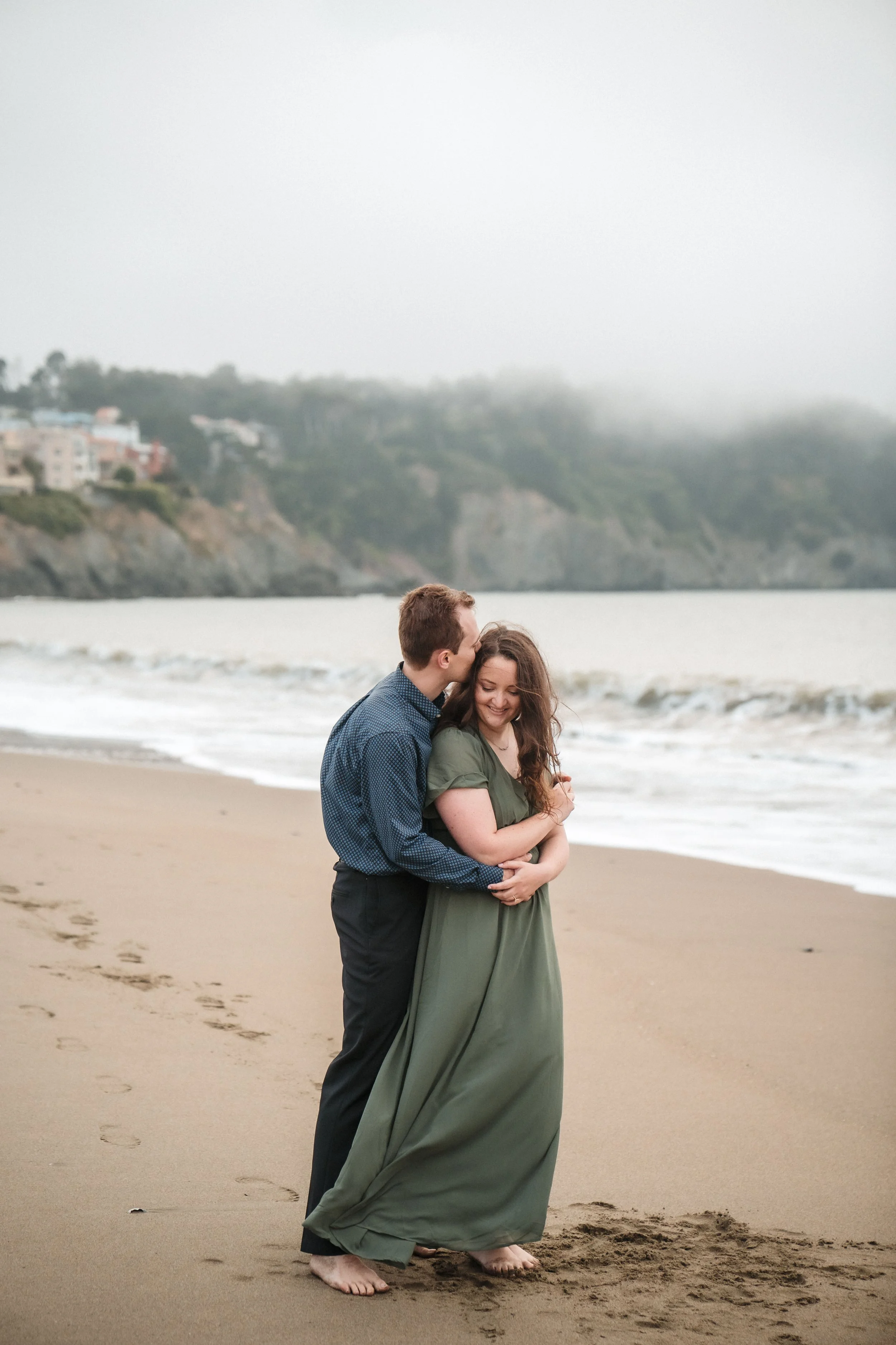 A couple standing close together on a sandy beach, hugging and smiling, with ocean waves and a hilly coastline in the background during their Bay Area engagement photos. Photography by Katherine Nicole Photography.