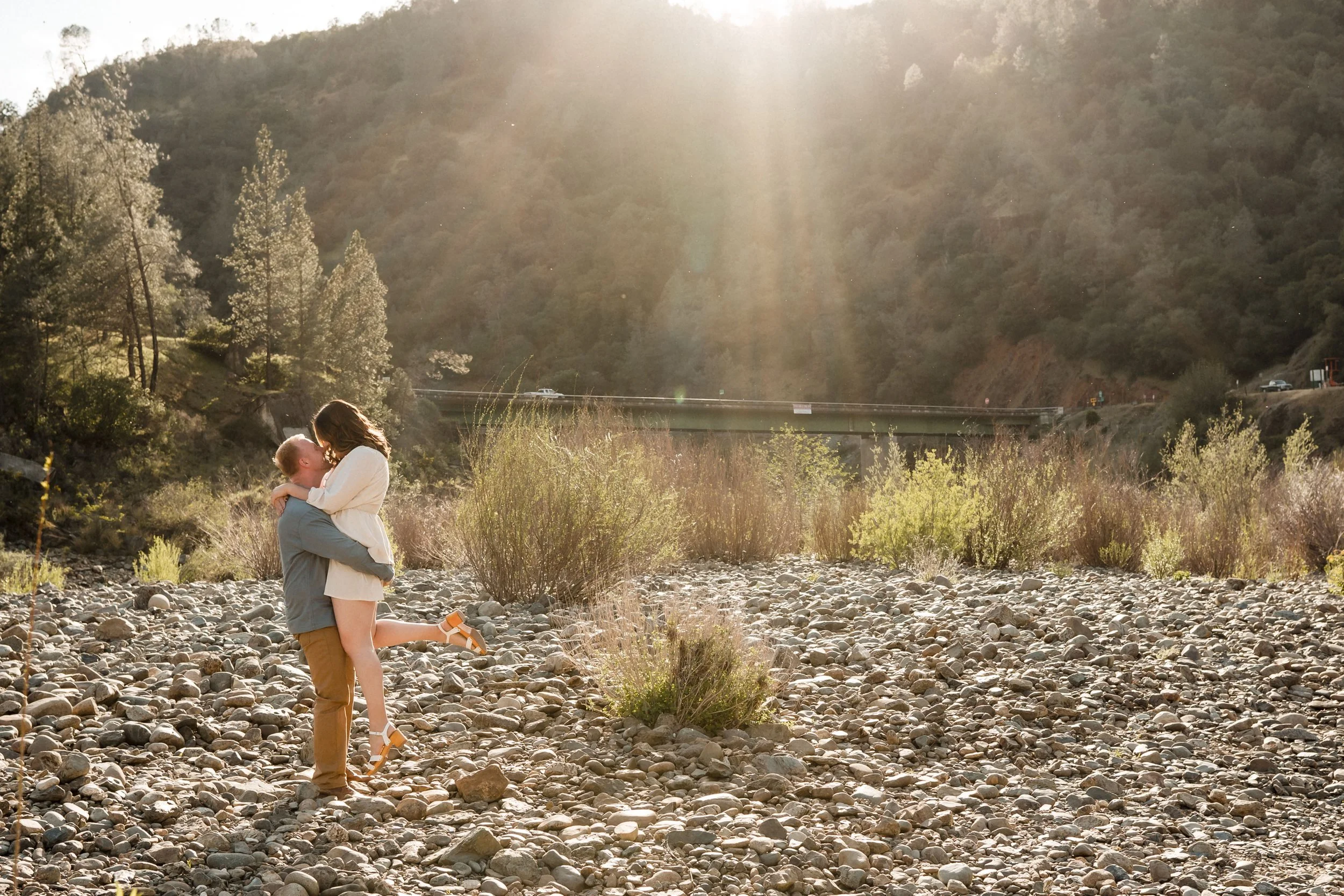 A couple sharing a kiss outdoors near the American River in Auburn during their engagement photos, with trees and hills in the background, illuminated by sunlight. Photography by Katherine Nicole Photography.