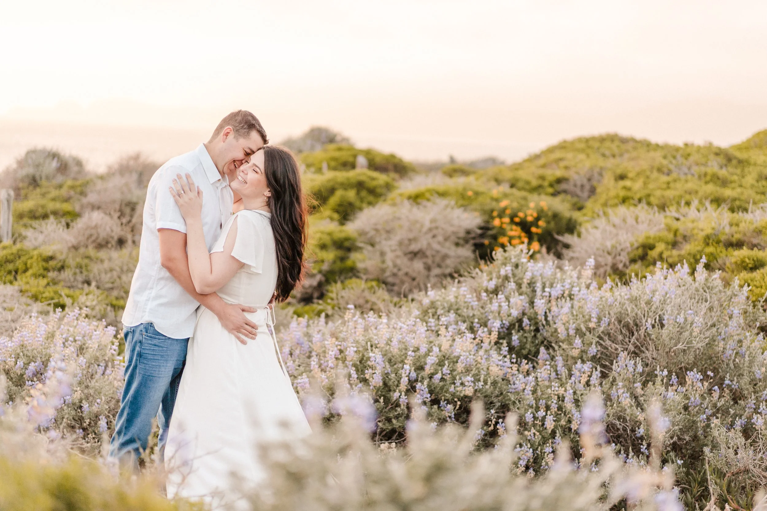A smiling couple embracing in a field of purple and white wildflowers during sunset while taking their engagement photos with Katherine Nicole Photography.