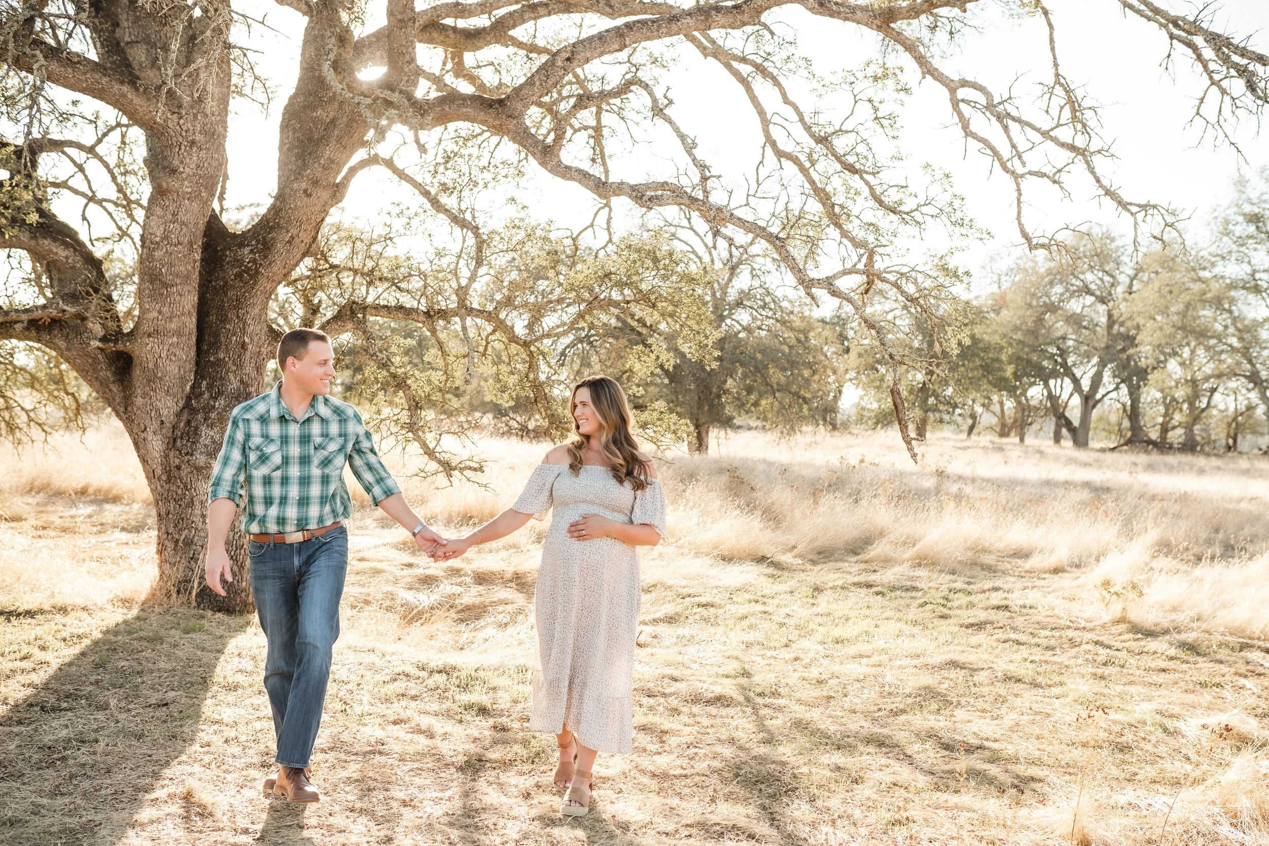 A smiling man in a green plaid shirt and jeans holding hands with a pregnant woman, walking outdoors under a large tree on a sunny day during a maternity session with Sacramento family photographer, Katherine Nicole Photography.