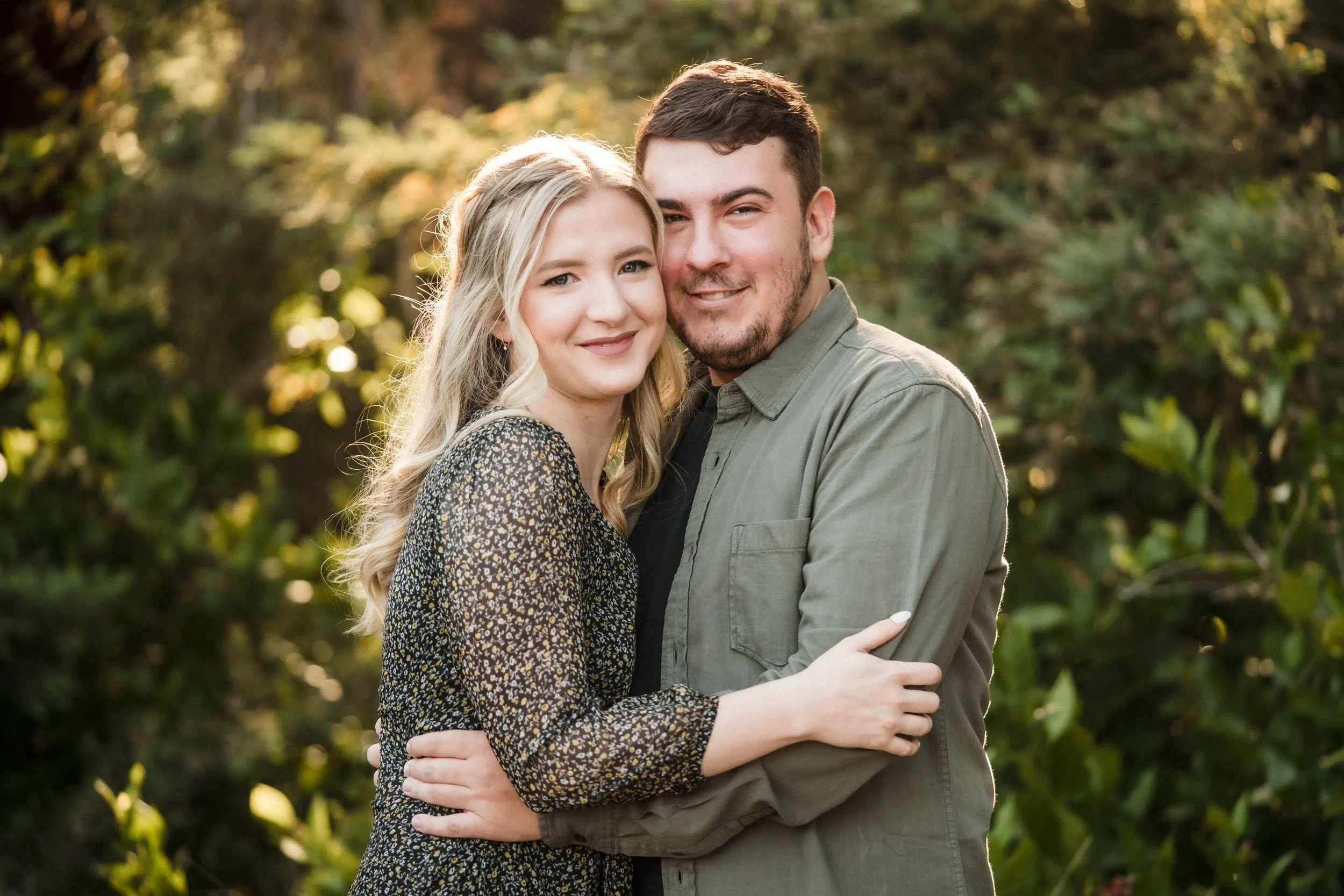 A young couple embracing outdoors in a natural setting with trees and greenery in the background during their engagement session. Photography by Katherine Nicole Photography.