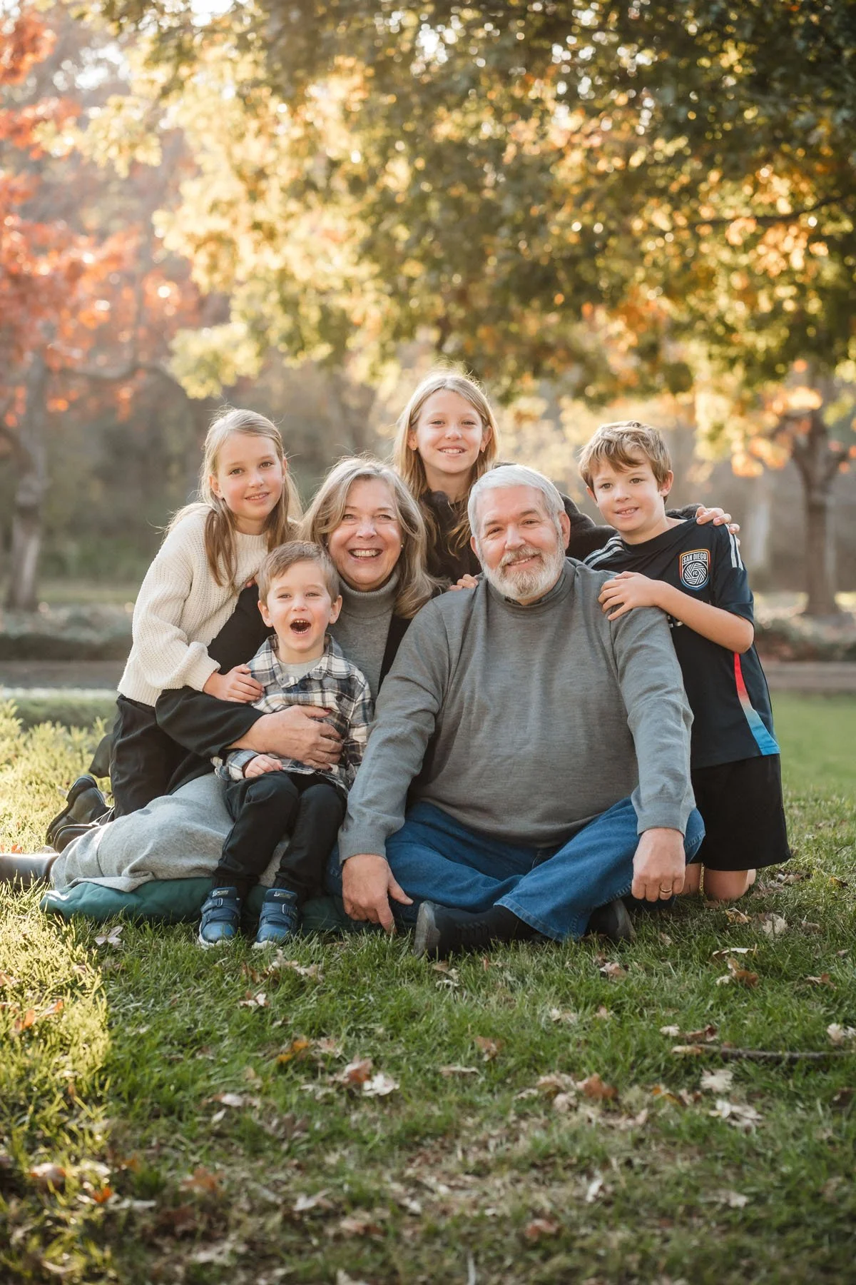 A multi-generational family of seven people sitting and kneeling on grass in a park during fall, smiling at the camera while taking family photos with Sacramento family photographer, Katherine Nicole Photography.