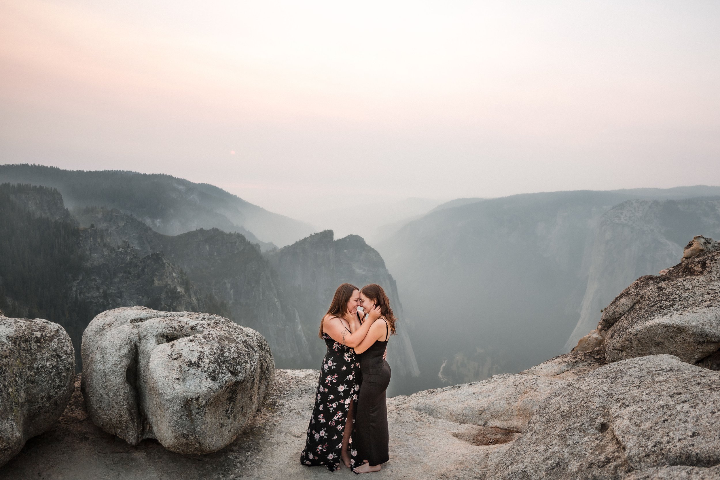 Two women sharing a moment at their engagement session at Taft Point in Yosemite, surrounded by large rocks and mountains in the background. Photography by Katherine Nicole Photography.