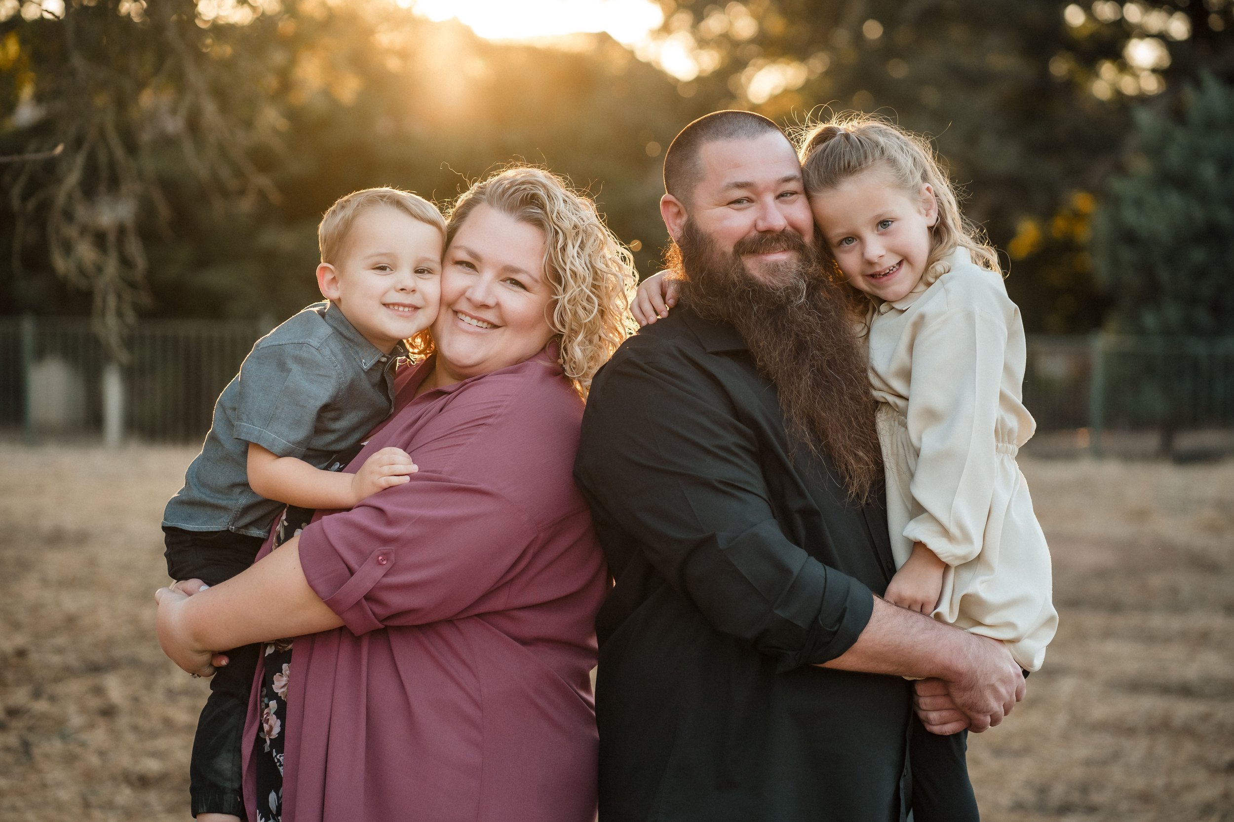 A family of four posing outdoors during sunset, with trees in the background, smiling and hugging each other during their family photos with Katherine Nicole Photography.