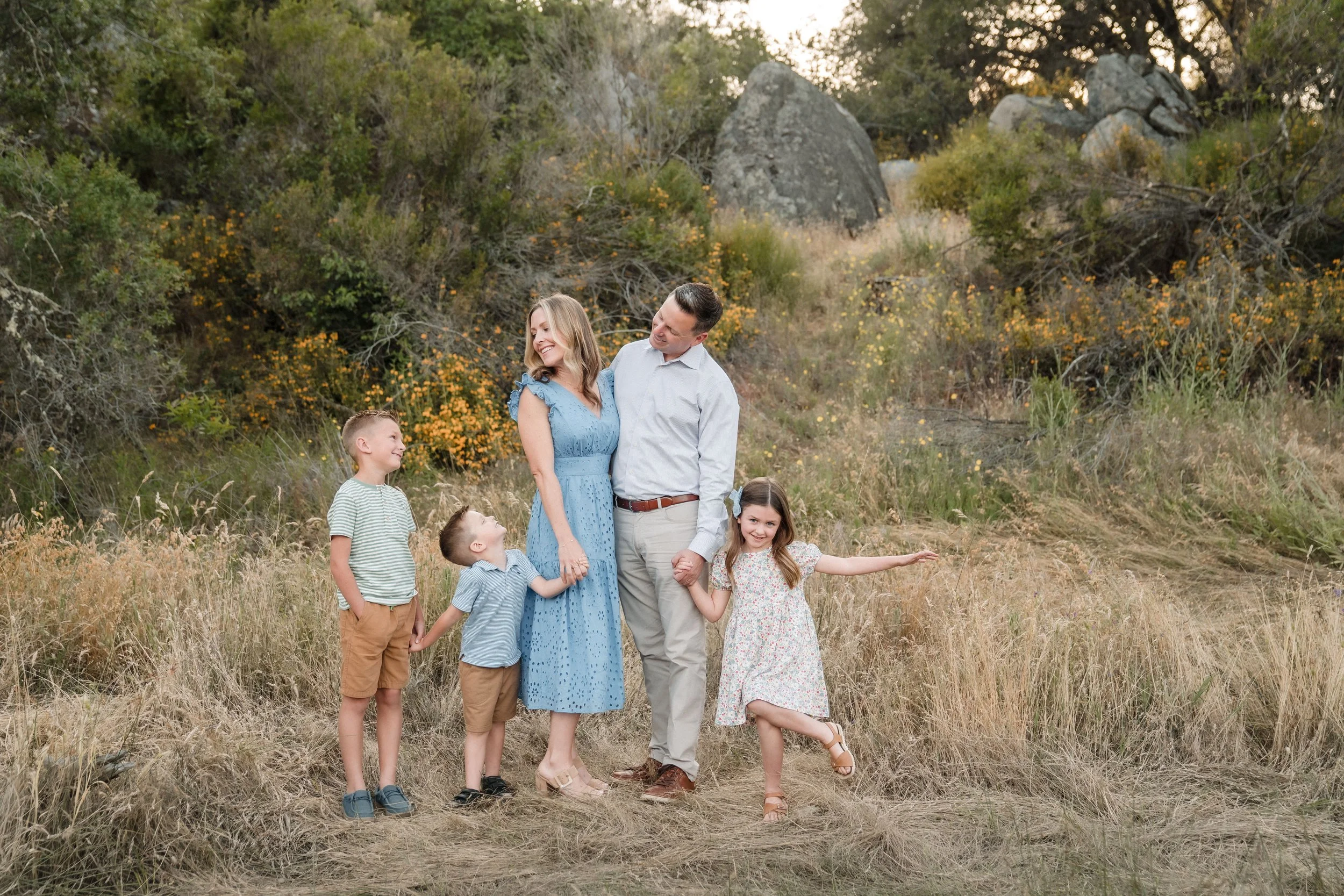 A family, standing together outdoors in a grassy, wooded area during sunset. The family appears to be happy, holding hands and smiling at each other during a family photo session with Sacramento family photographer Katherine Nicole Photography. 