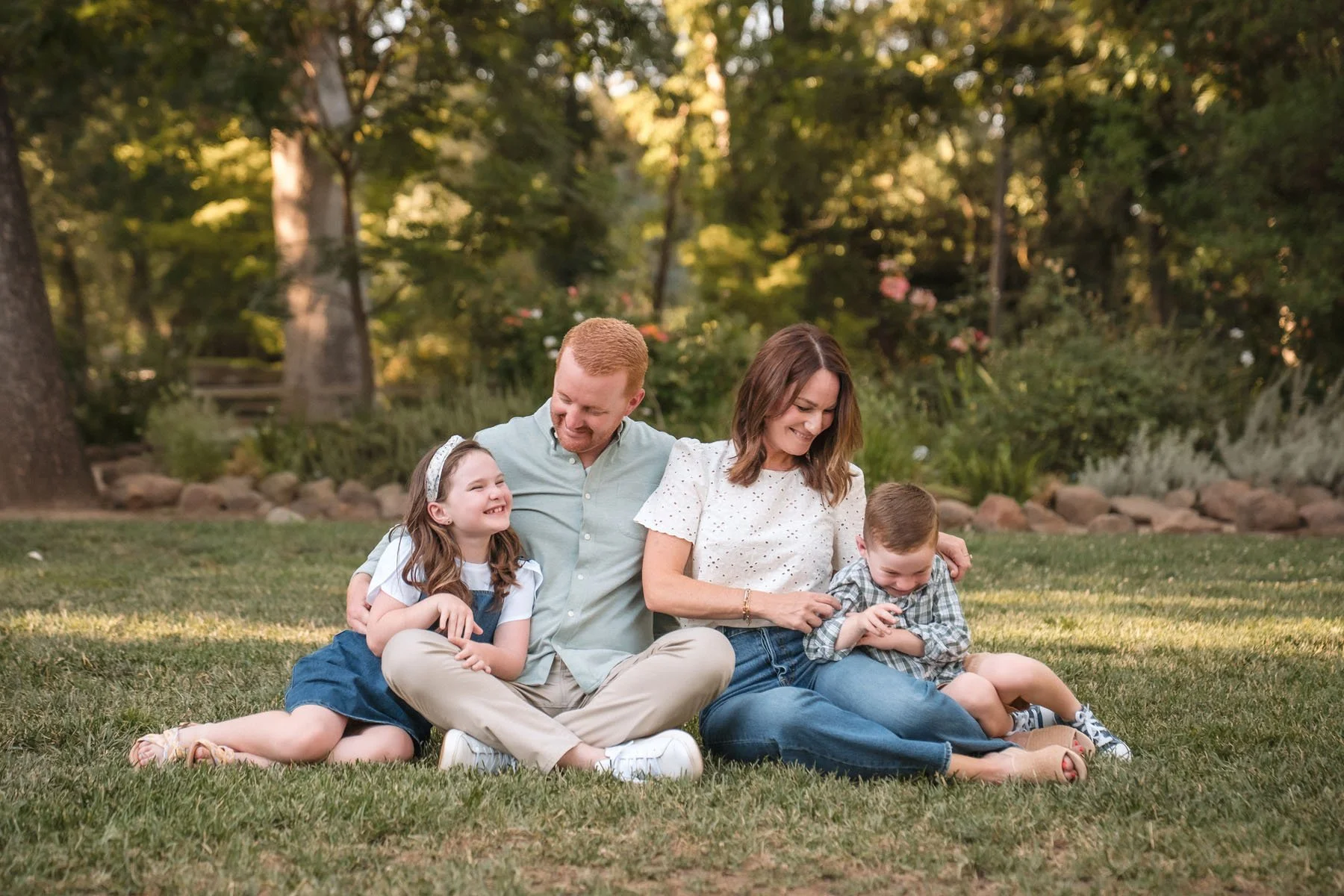A family of four sitting on grass in a park, smiling and sharing a joyful moment during the evening, with trees and rocks in the background.