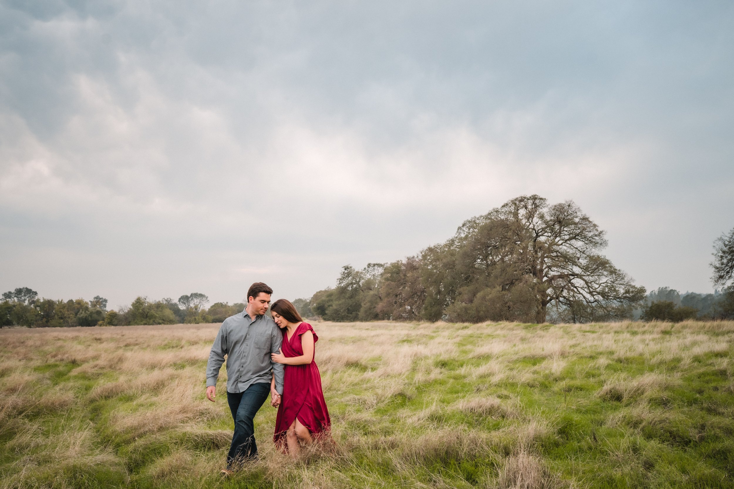 A young couple holding hands and standing close together in a grassy field with large trees and an overcast sky in the background, during their Folsom engagement session with Katherine Nicole Photography.