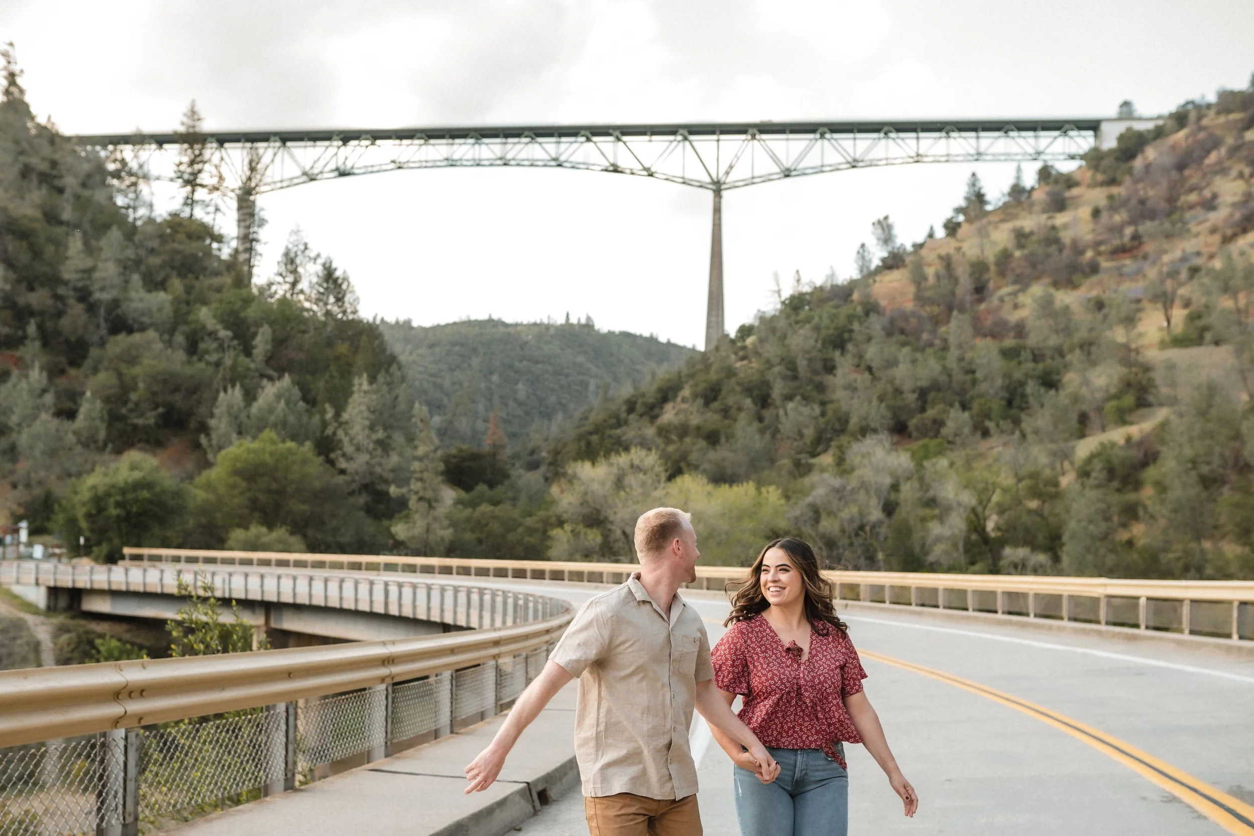 A couple walking hand in hand with the Foresthill Bridge and a mountainous landscape in the background, during their engagement session with Katherine Nicole Photography.