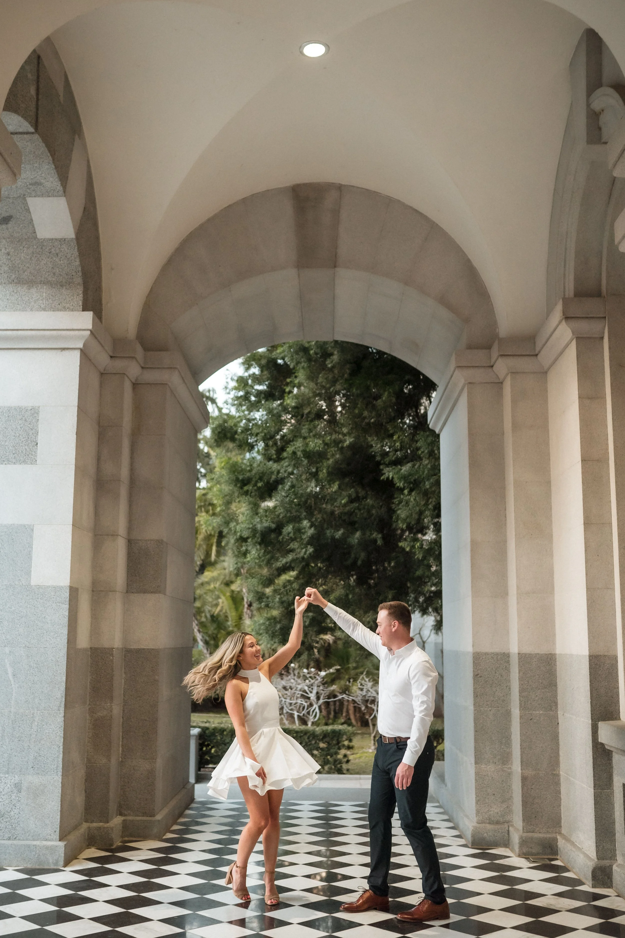 A couple dancing at the Sacramento Capitol building, trees in the background, the woman in a white dress, and the man in a white shirt and dark pants, holding hands and smiling, while taking their engagement photos with Katherine Nicole Photography.