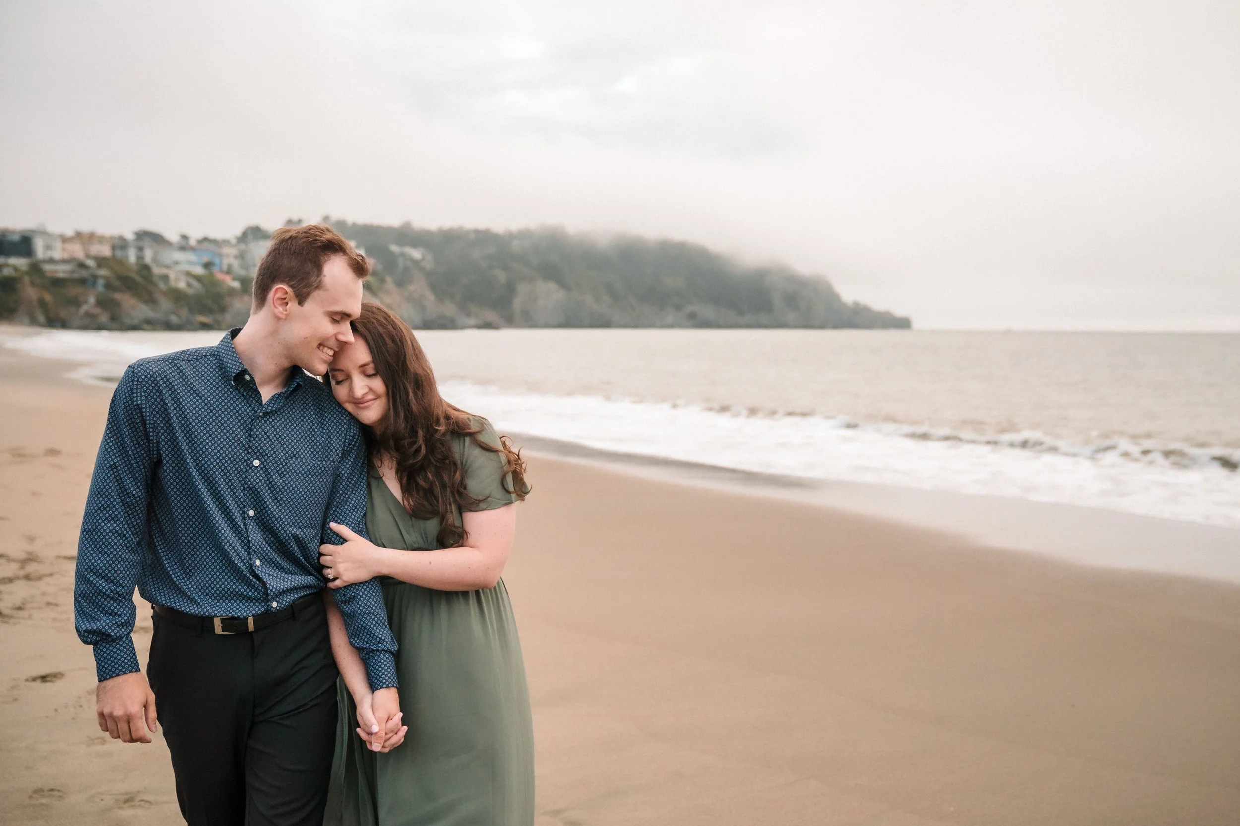 A young couple stands on Baker Beach during their engagement session, lovingly embracing each other with their eyes closed, a foggy coastline and houses visible in the distance under a cloudy sky. Photography by Katherine Nicole Photography.