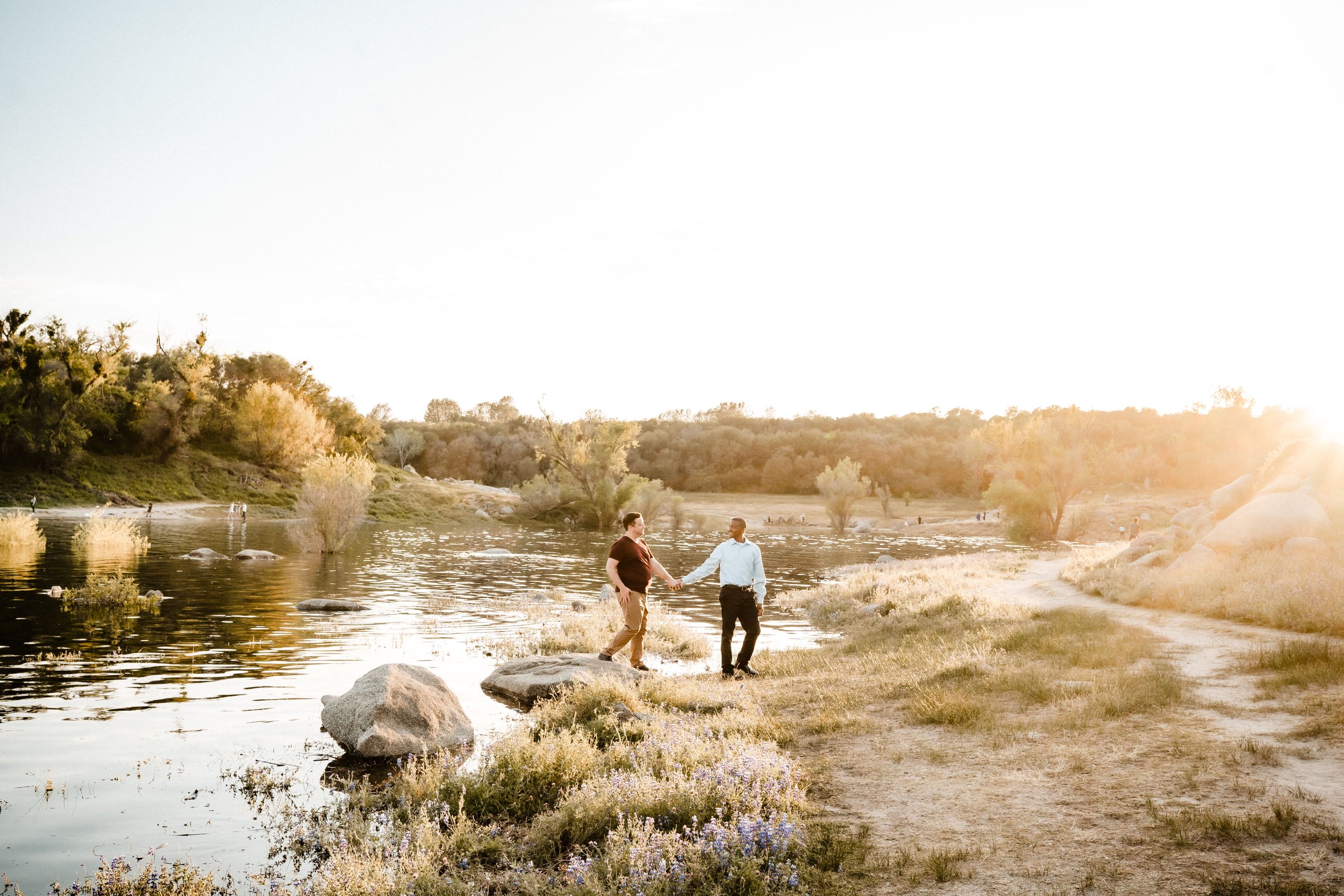 Two men holding hands near Folsom Lake at sunset, in a natural setting with trees and rocks, during their engagement session with Katherine Nicole Photography.