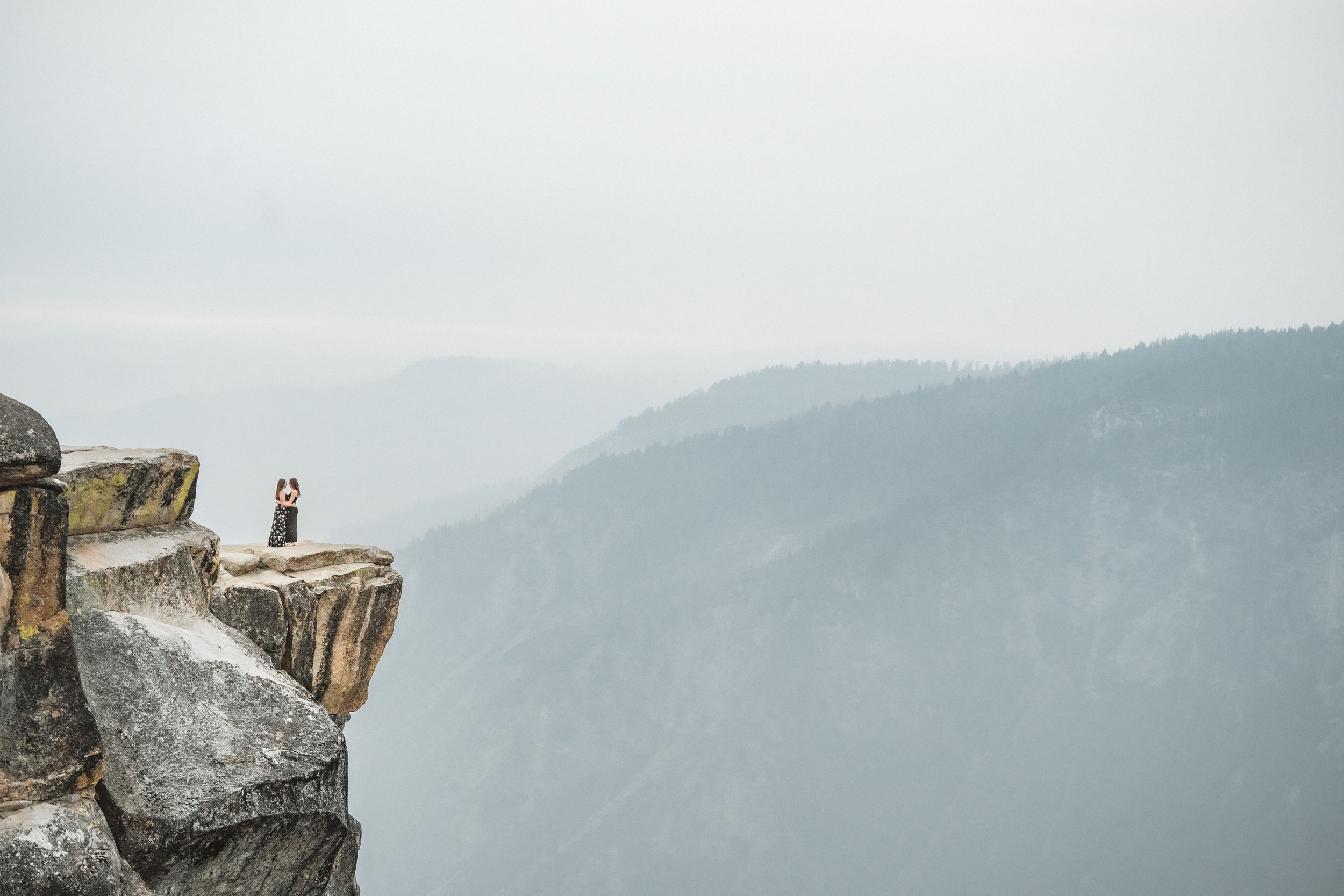 A couple embracing on a rocky ledge at Taft Point in Yosemite, during their engagement session overlooking a misty mountain landscape. Photography by Katherine Nicole Photography.