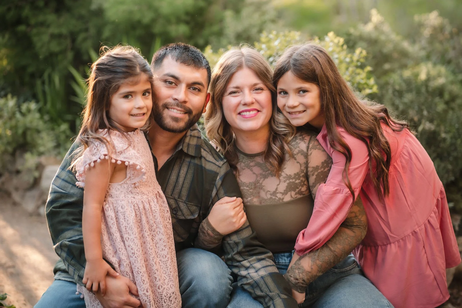 A happy family of four, with two young girls and their parents, smiling and posing outdoors in a natural setting with greenery, taking family photos with Katherine Nicole Photography.