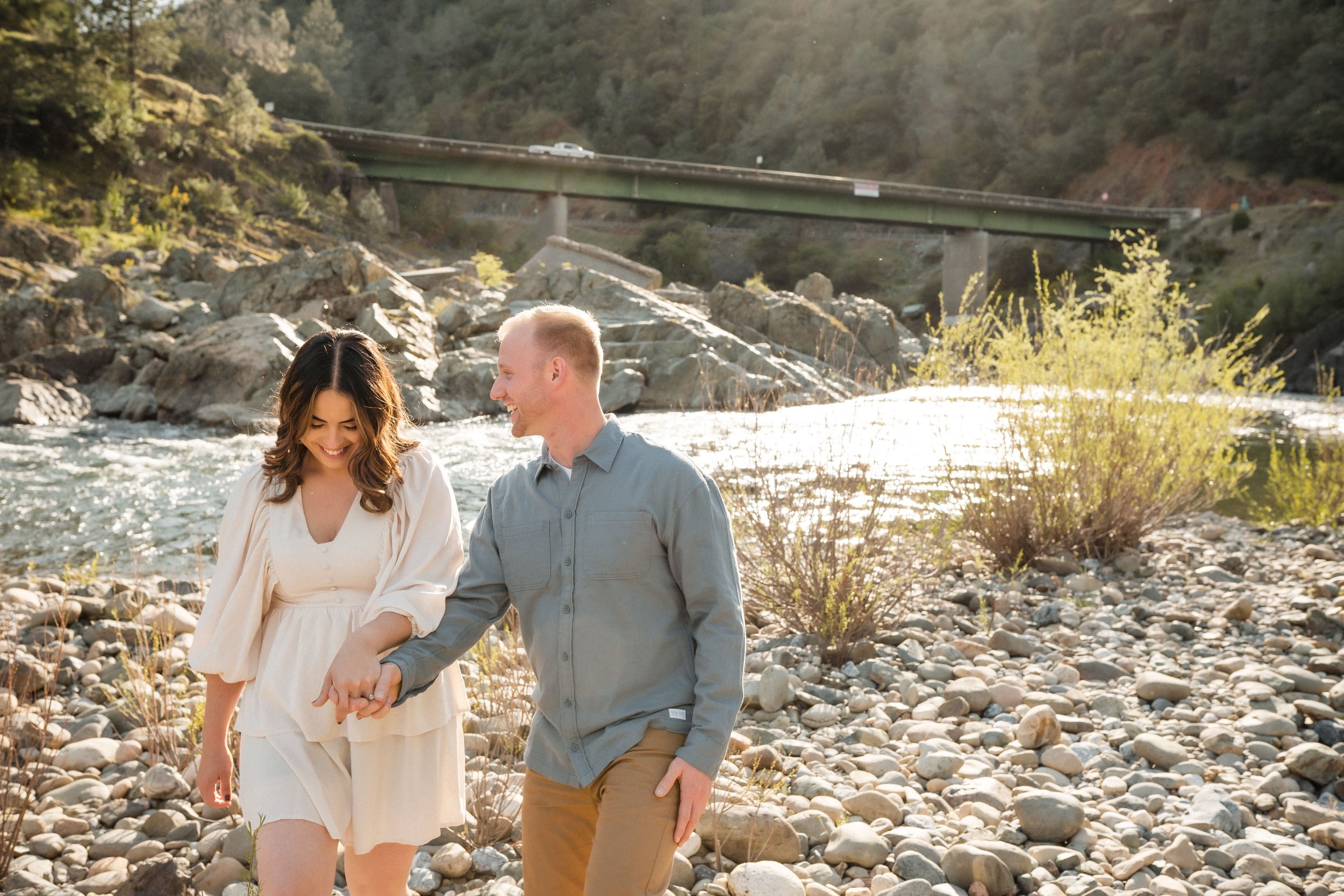 A smiling couple holding hands and walking along the American River in Auburn, with water, trees, and a bridge in the background, while taking their engagement photos with Katherine Nicole Photography.