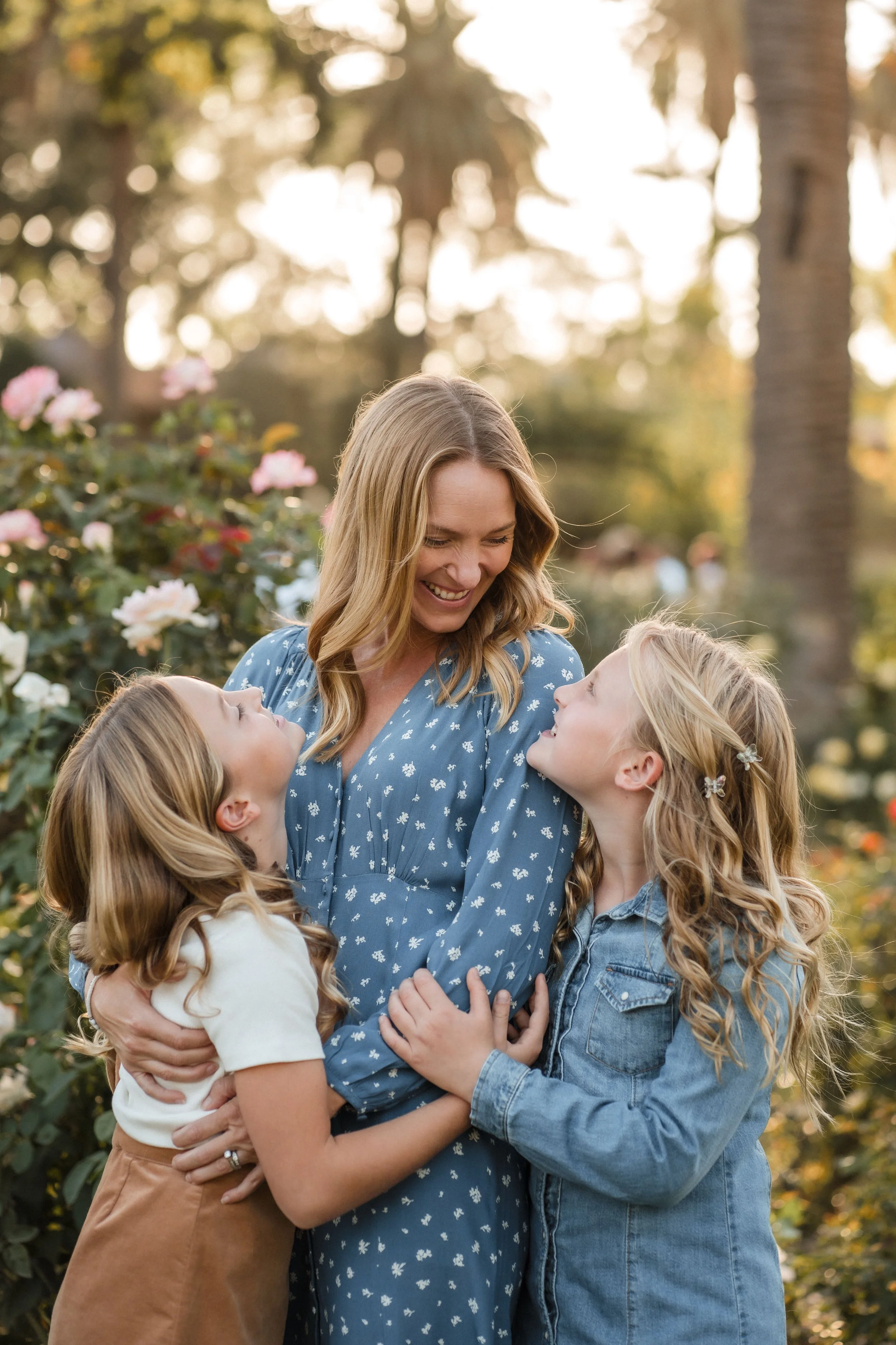 A woman and two young girls are embracing outdoors, surrounded by plants and flowers, with sunlight filtering through trees in the background while they take family photos at the McKinley Park rose garden with Katherine Nicole Photography.