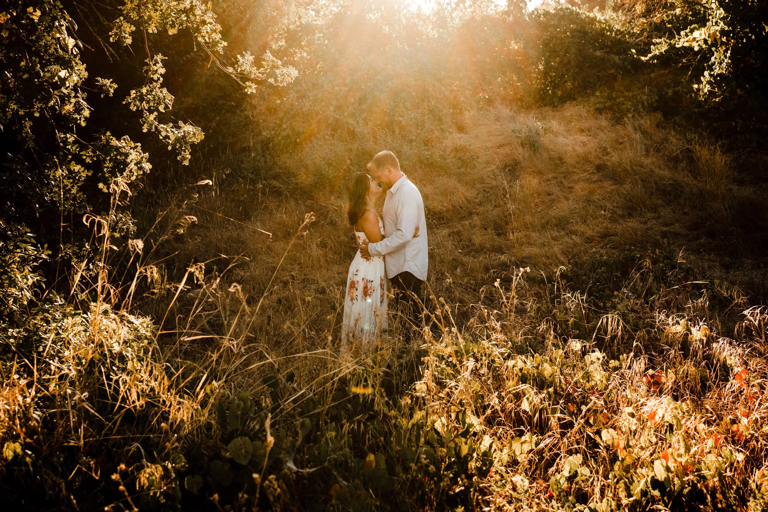 A couple standing close together in a grassy outdoor area with sunlight shining down, embracing and looking at each other, during their Sacramento engagement session with Katherine Nicole Photography.