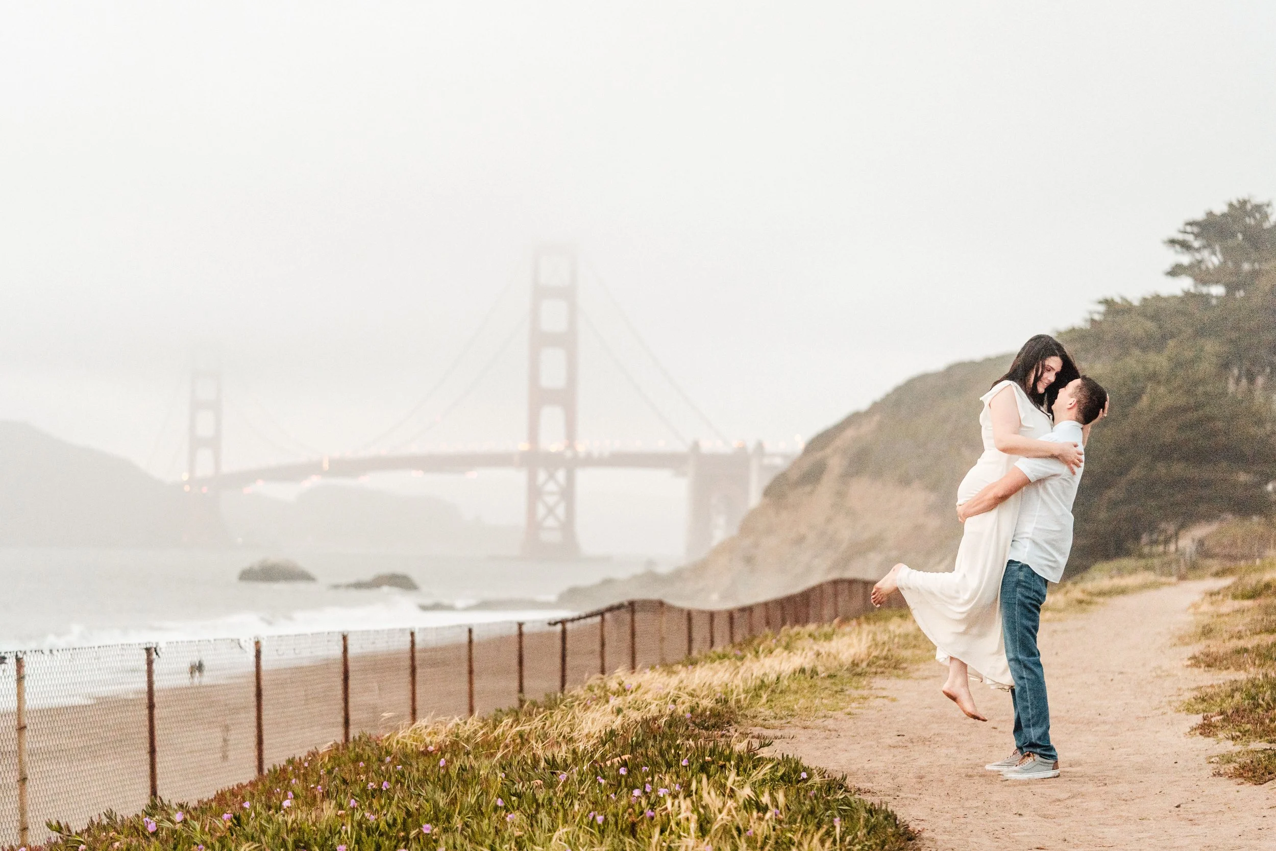 A couple enjoying a moment together near Baker Beach with the Golden Gate Bridge in the background. The man is lifting the woman, and they are both smiling, with fog partially obscuring the bridge. Photography by Katherine Nicole Photography.