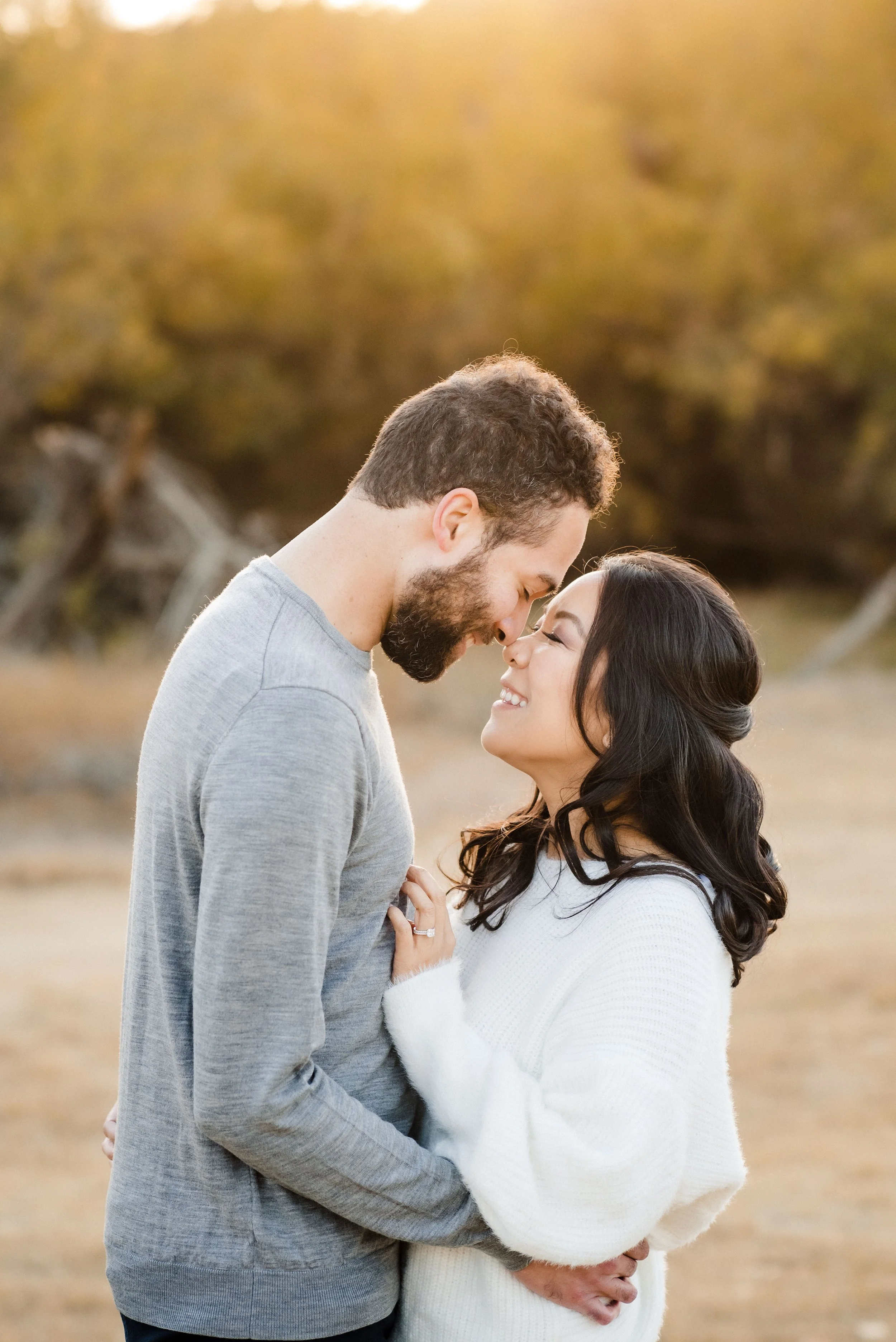 A couple sharing an intimate moment during their engagement photos, standing close with foreheads touching and smiling at each other, with a blurred background of trees and golden sunlight. Photography by Katherine Nicole Photography.
