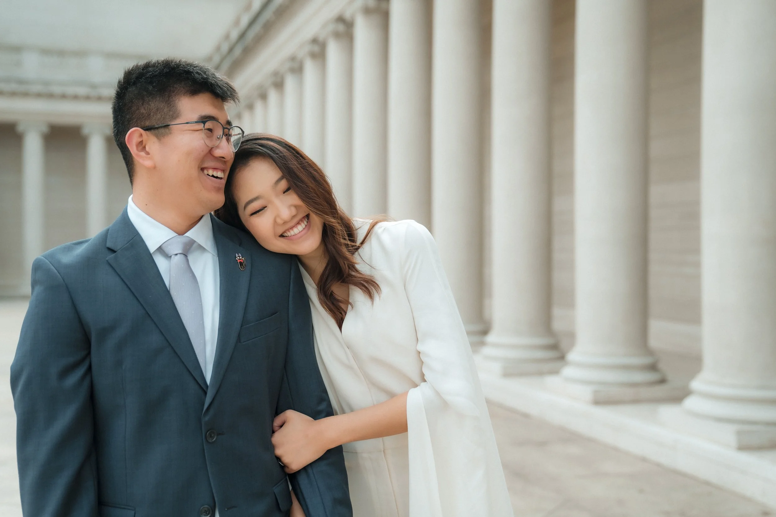 A smiling man in a gray suit and glasses hugging a smiling woman during their engagement photos at the Legion of Honor in San Francisco. Photography by Katherine Nicole Photography.