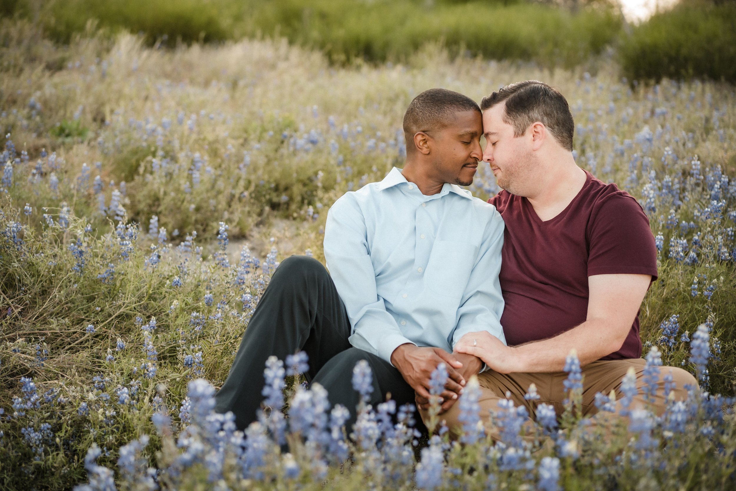Two men sitting close together in a field of blue flowers with their foreheads touching and eyes closed, sharing an intimate moment during their engagement session at Folsom Lake. Photography by Katherine Nicole Photography.